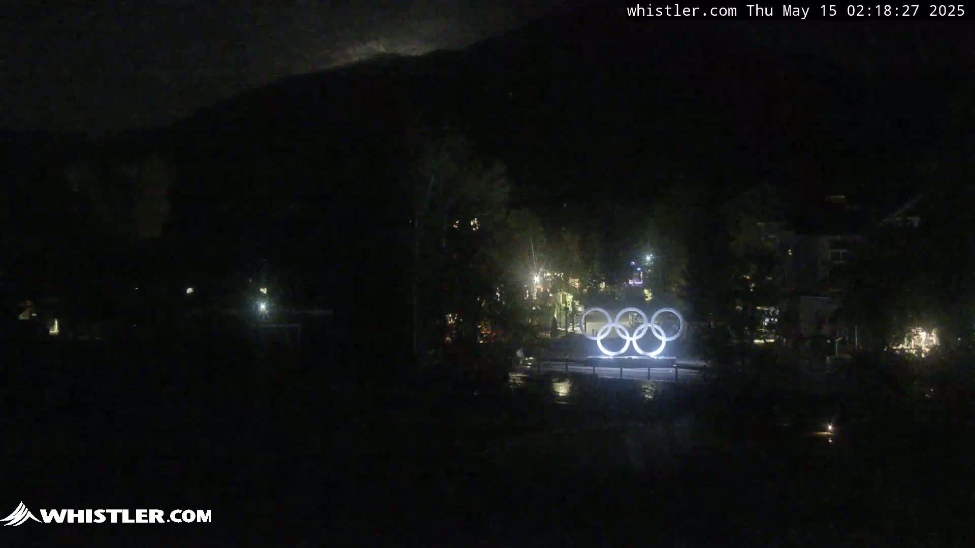 A nighttime view of the Olympic rings illuminated in front of a dark, mountainous landscape under a partly cloudy sky.