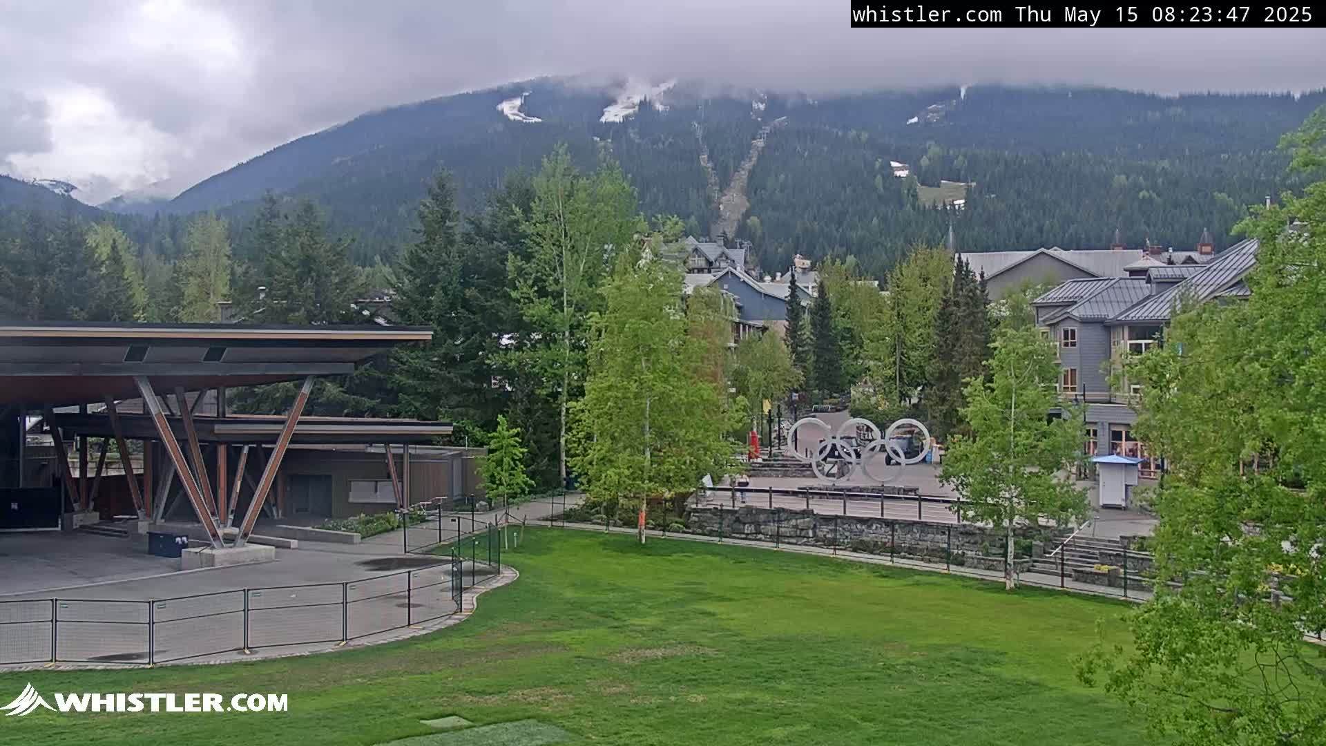 A view of a grassy area with a modern building, trees, and a mountain range in the background under cloudy skies.