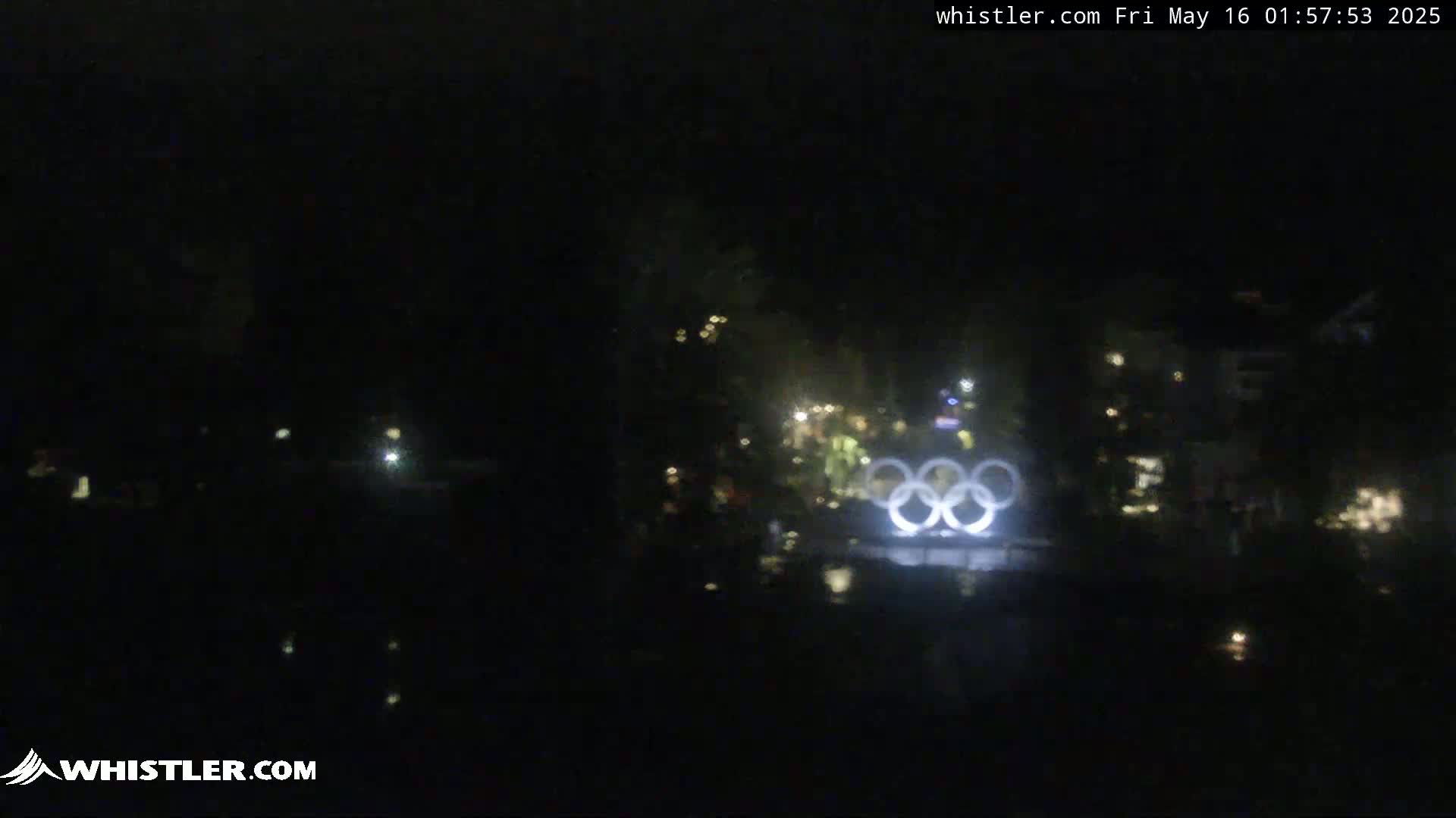 At night, illuminated Olympic rings are visible in the distance, surrounded by dark trees and faintly lit buildings.