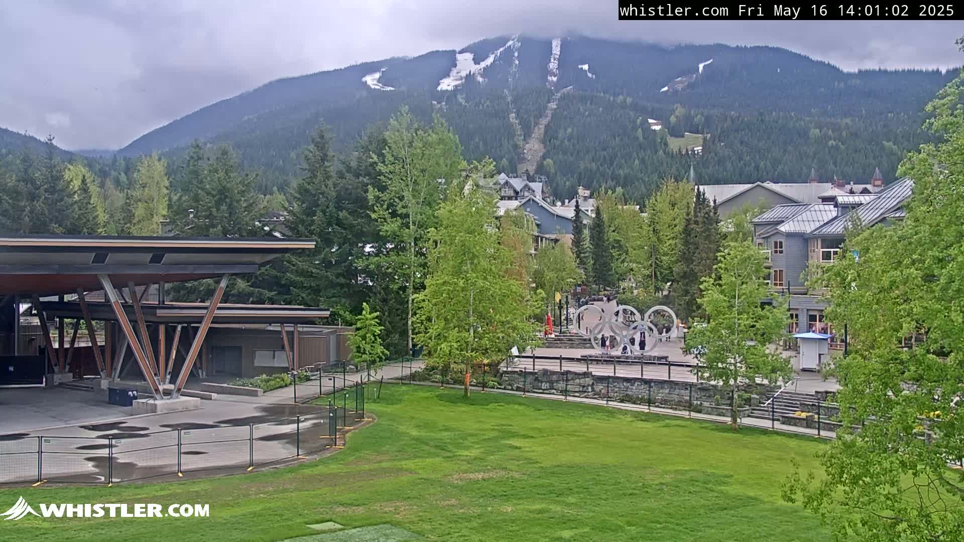 A mountain village under cloudy skies, featuring a grassy area, a modern building with a large canopy, and a mountain range in the background with some remaining snow patches.