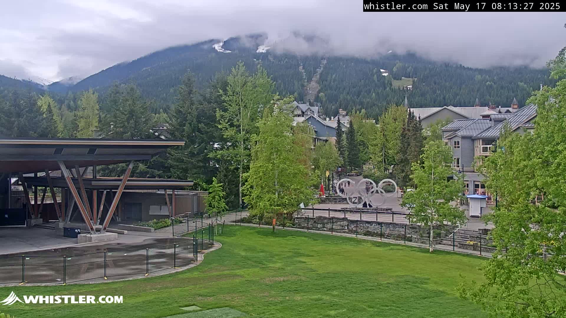 A partially cloudy day reveals a mountain range partially obscured by clouds behind a village of buildings, a grassy field, and a modern structure with a peaked roof.