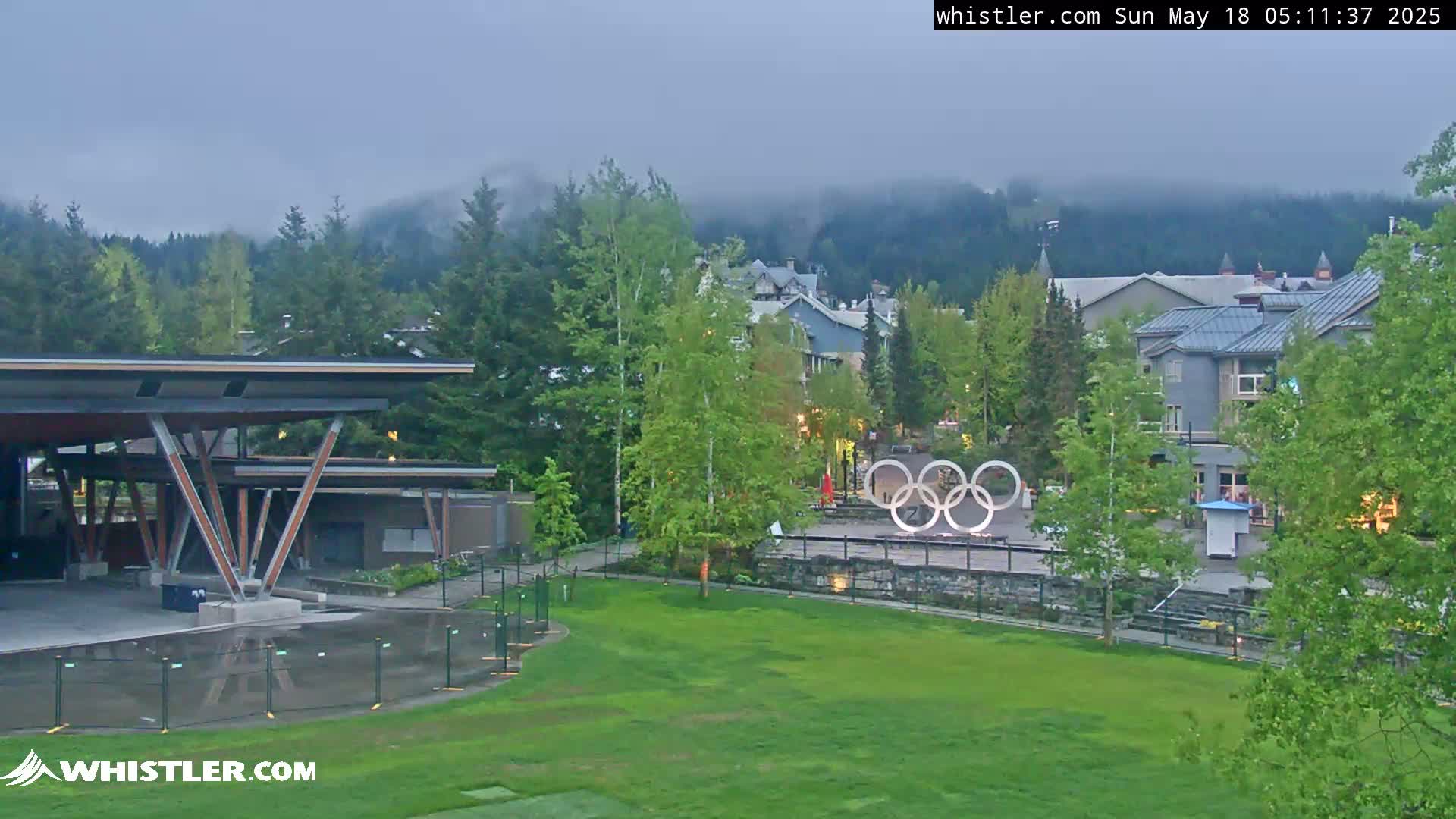 A view of a grassy area with a modern pavilion, Olympic rings, and buildings nestled amongst lush green trees under an overcast sky.