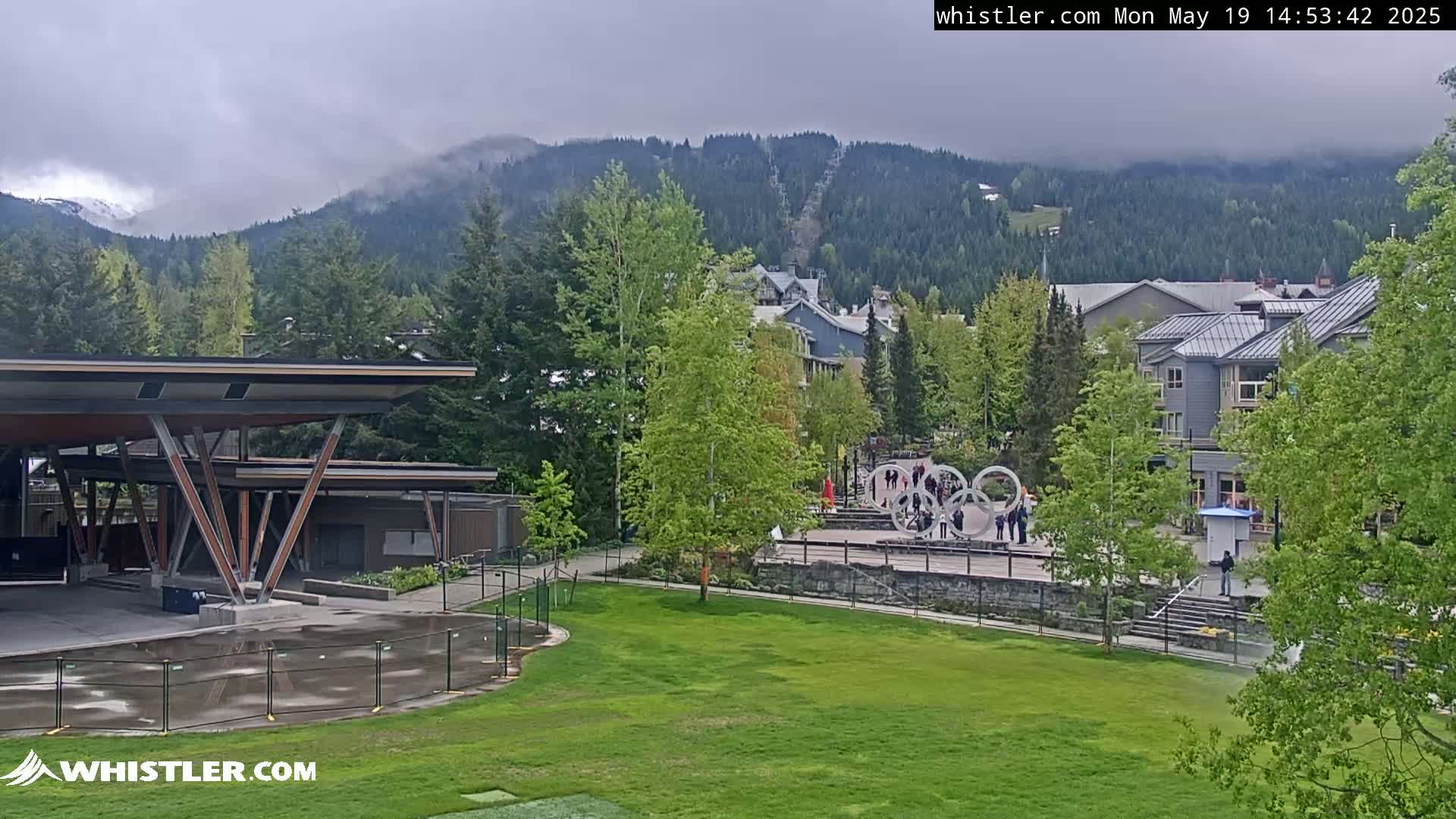 A grassy area with a modern pavilion overlooks a village nestled in a valley, with overcast skies and mountains in the background partially obscured by mist.