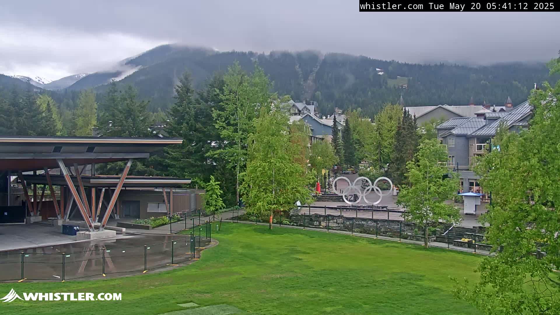 A partially cloudy day reveals a mountain range partially obscured by fog, overlooking a village with green trees, buildings, and an Olympic rings monument in a grassy area.