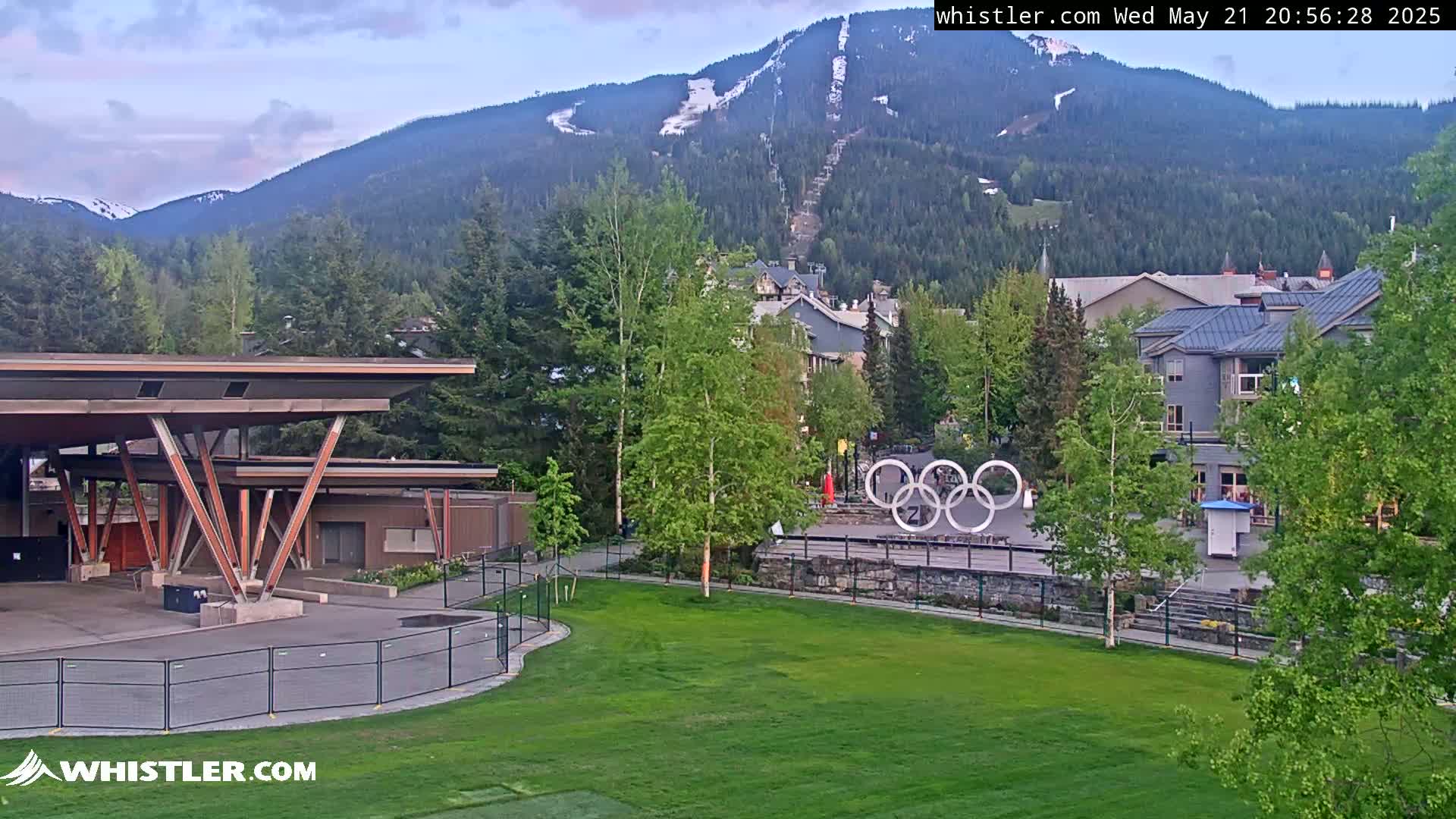 A grassy area with the Olympic rings in the background, situated below a mountain with some snow visible, under a partly cloudy sky.