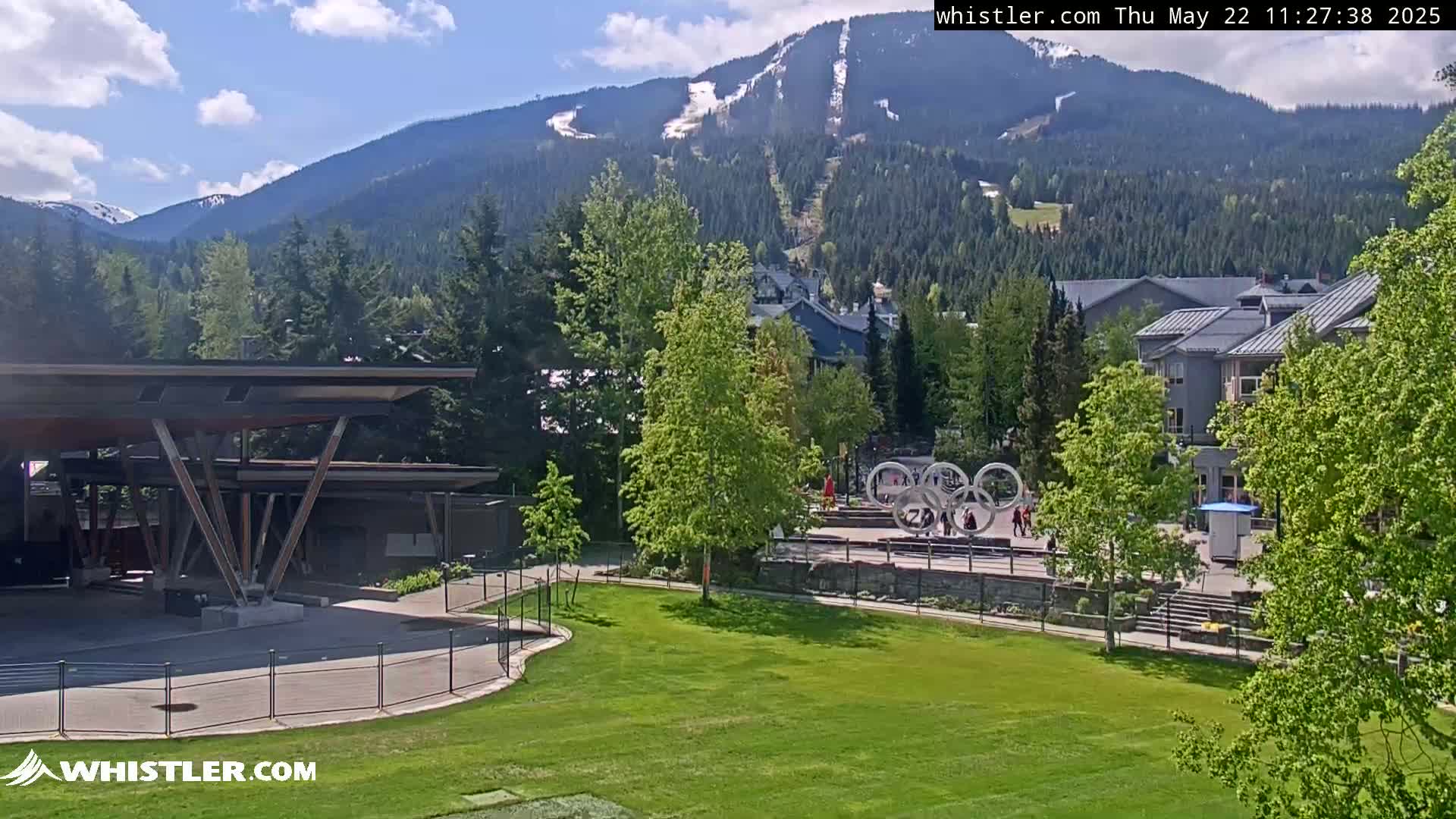 A partly sunny day reveals a mountainside ski resort with some remaining snow, overlooking a grassy area with a modern structure and Olympic rings sculpture.