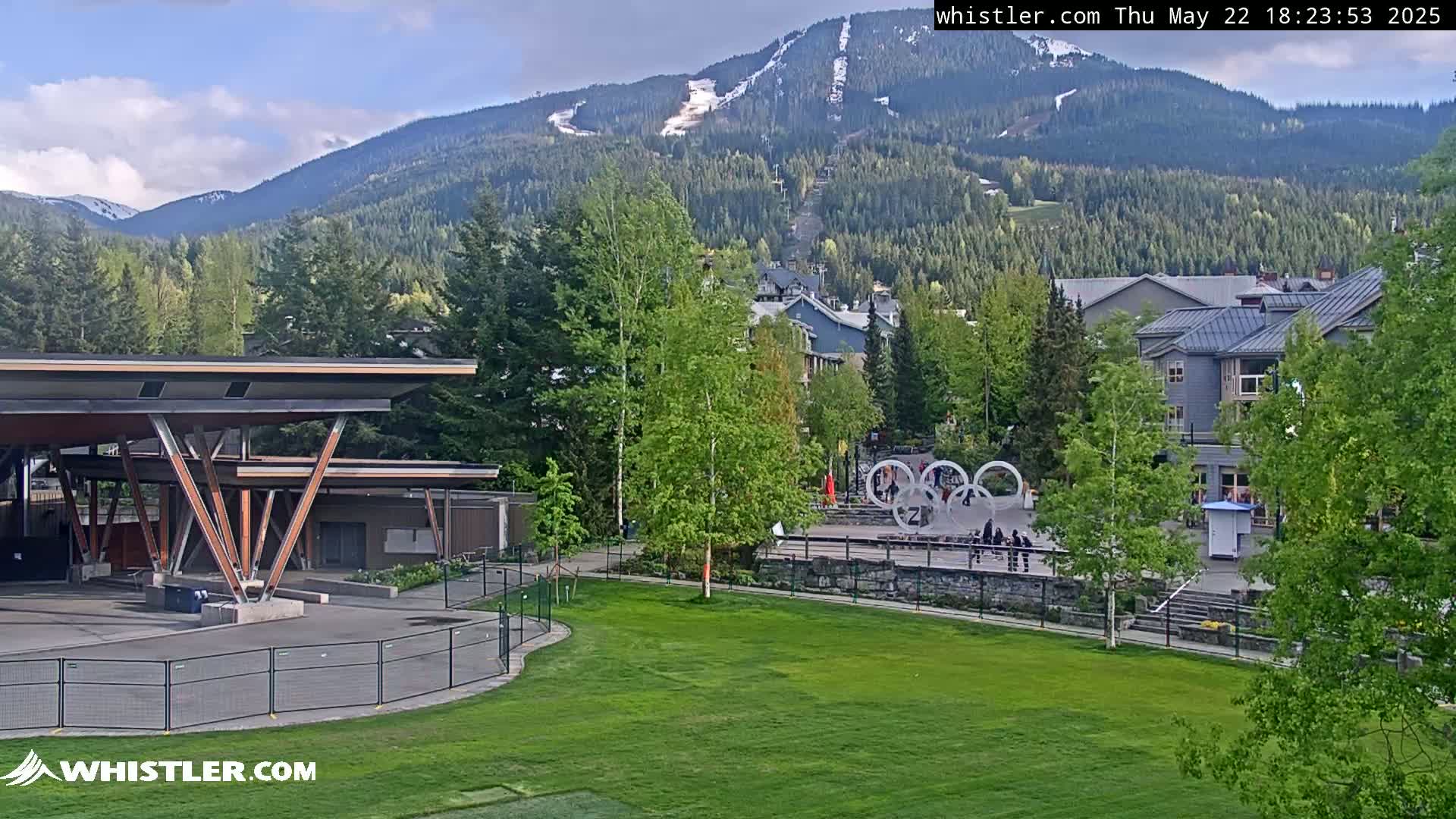 A partly cloudy day reveals a mountain village nestled in a valley, with a grassy park, modern buildings, and a snow-capped mountain in the background.