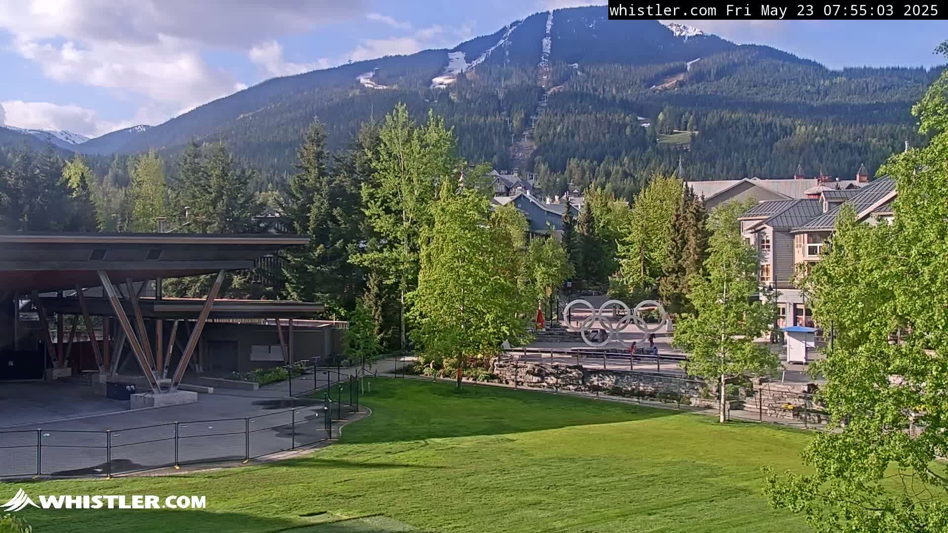 A partially sunny view of a grassy park area, with buildings, trees, and mountains in the background, some of which still have snow.