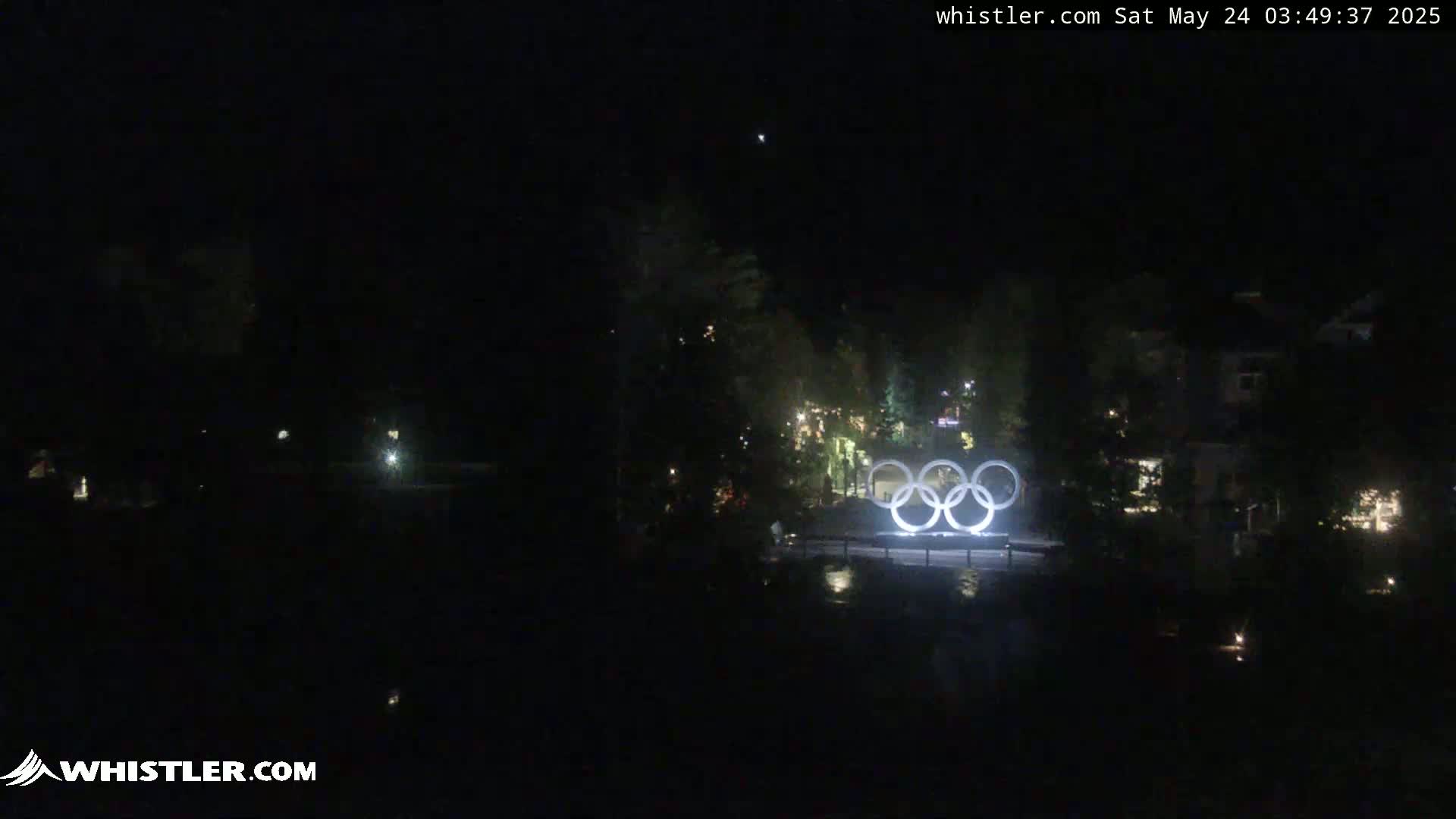 At night, illuminated Olympic rings are visible in the distance, with some dimly lit buildings and trees surrounding them.