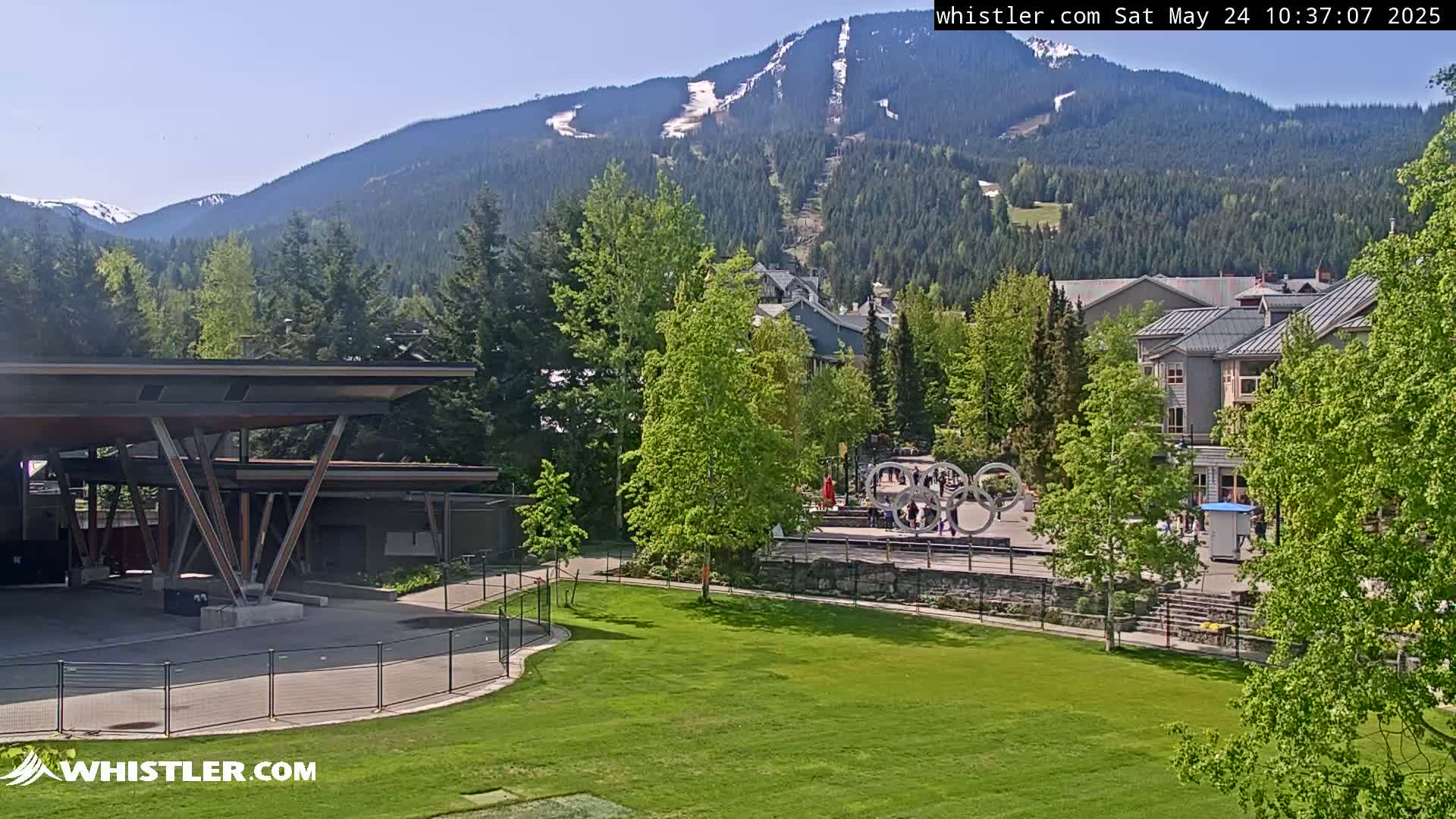 A sunny view of a grassy area with a modern structure in the foreground, some buildings and a playground in the midground, and a mountain range with a ski slope visible in the background.