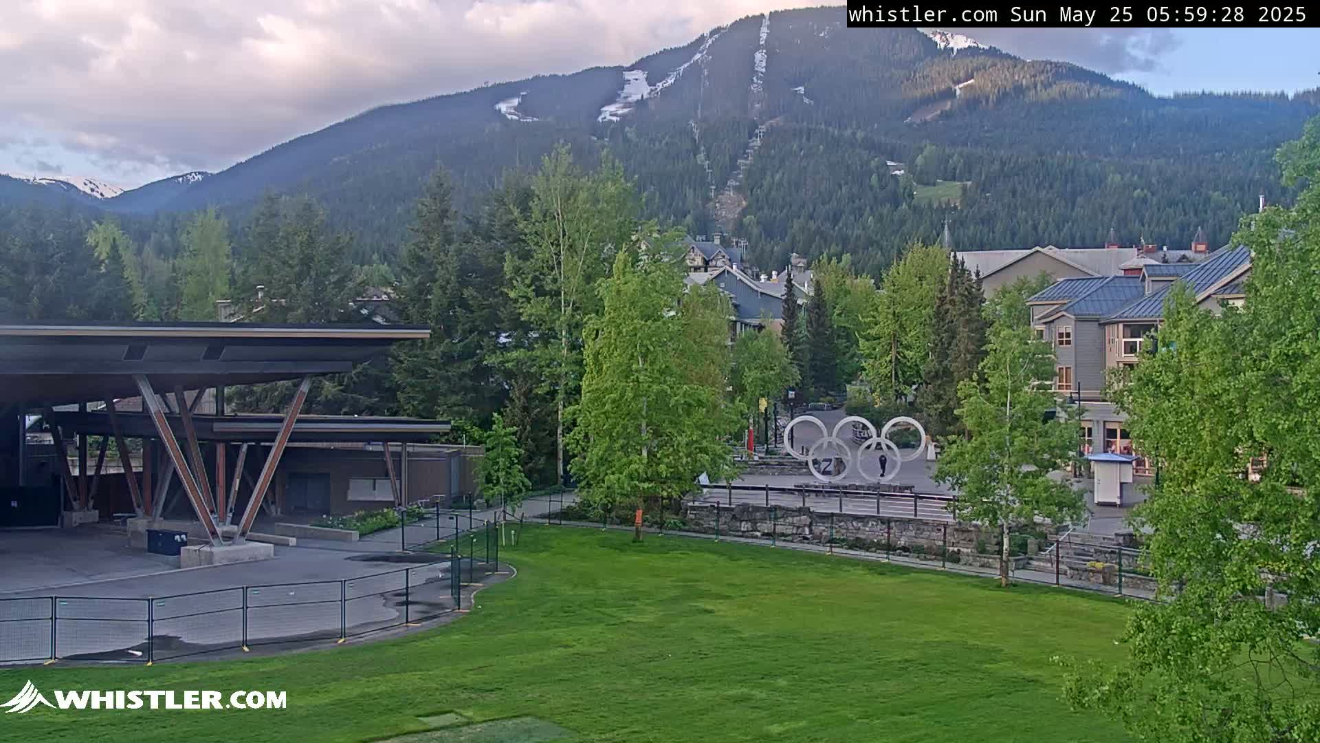 A grassy area with a modern pavilion and the Olympic rings sits in front of buildings and trees at the base of a mountain range under a partly cloudy sky.