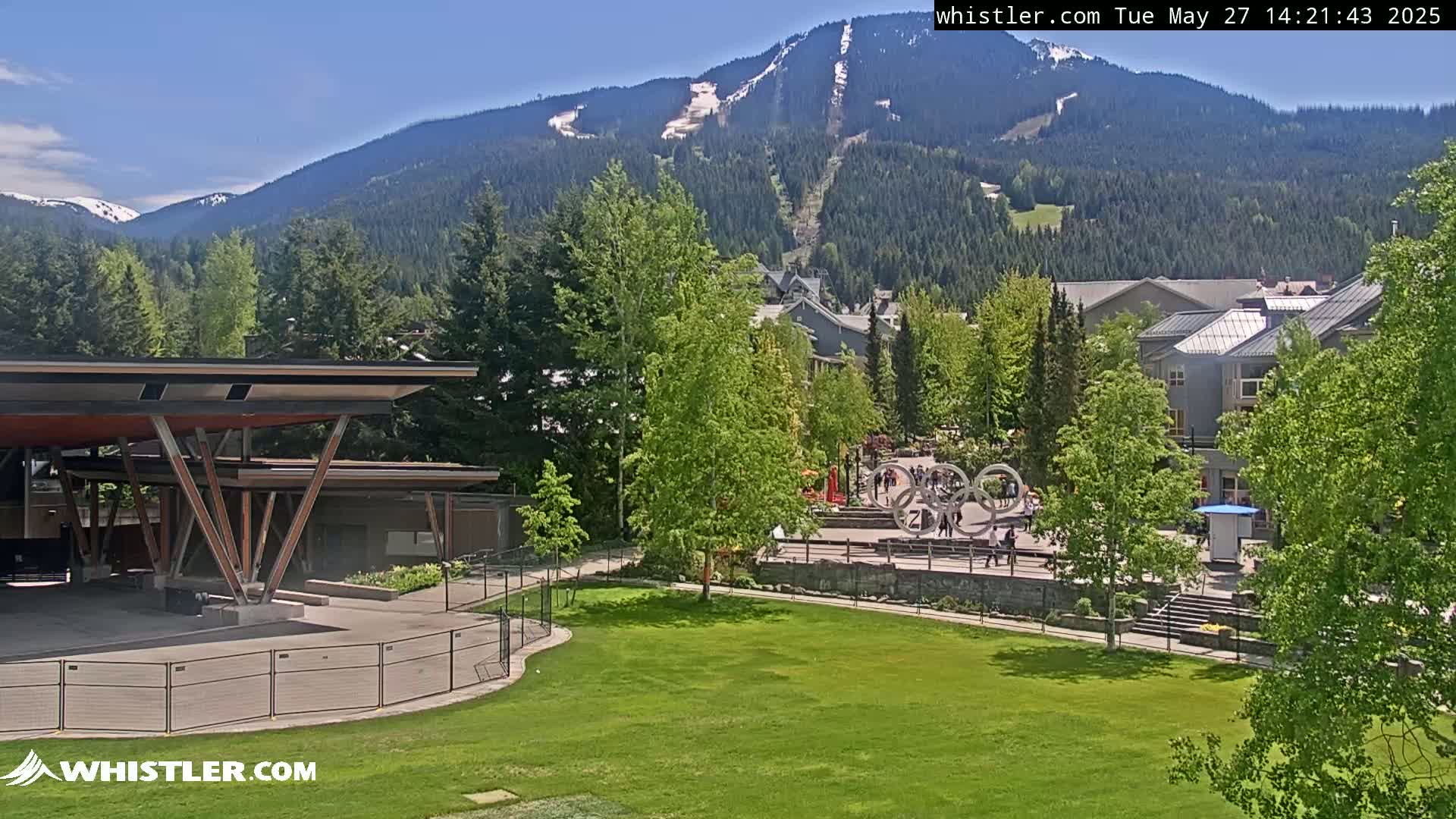 A grassy park area with a covered stage and surrounding buildings, under a sunny sky, is set against a backdrop of a mountain range with some remaining snow on its peaks.