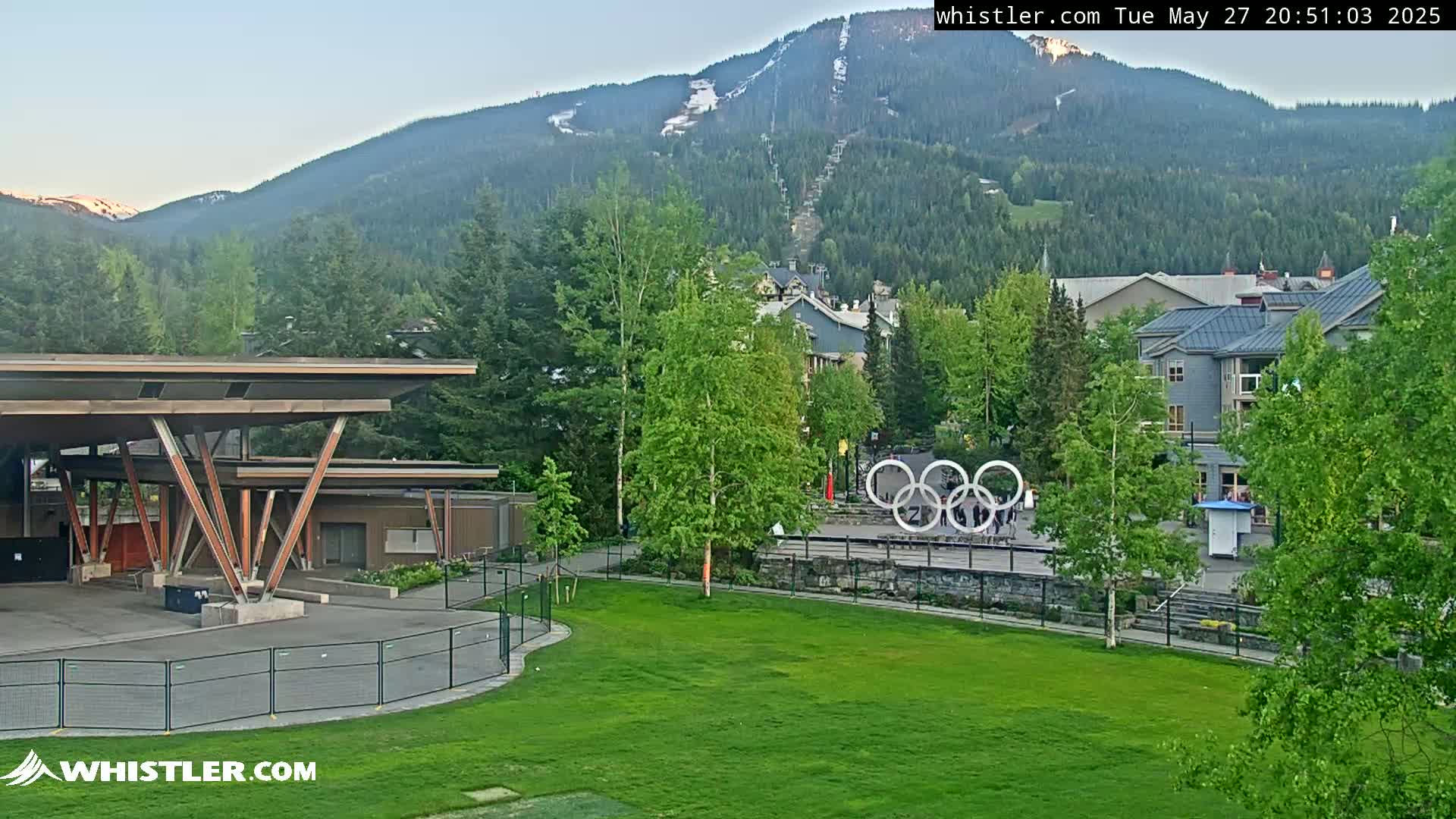 A grassy area with a modern covered structure, Olympic rings, and buildings sits in front of a mountain with a ski lift visible under a clear evening sky.