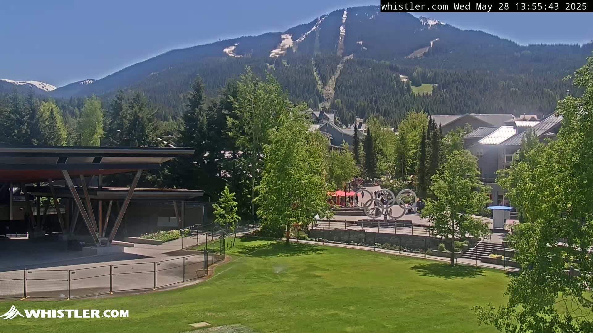 A sunny view of a grassy park with a modern pavilion, a mountain range in the background with some remaining snow, and a village nestled in the trees.