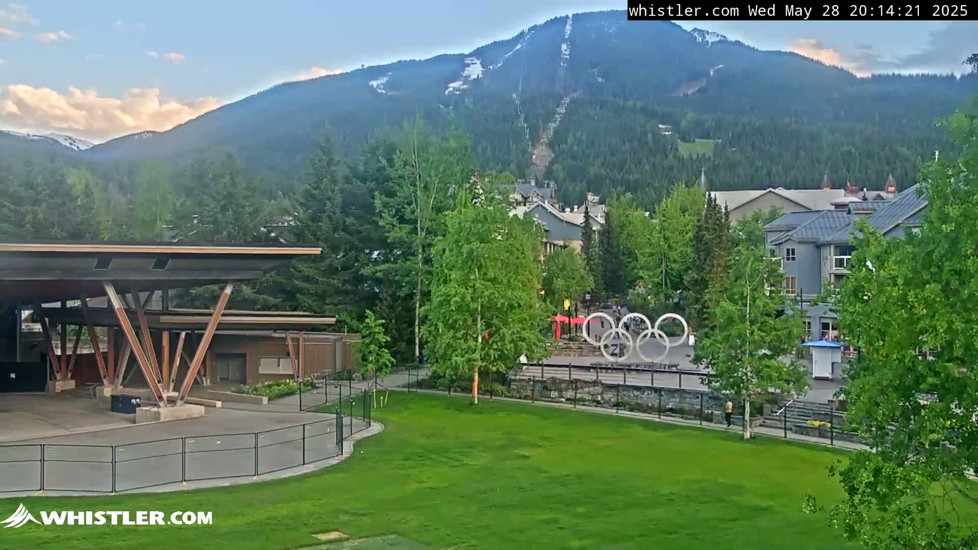 A mostly clear, late afternoon view of a mountainside ski resort town with a grassy park featuring the Olympic rings in the foreground.