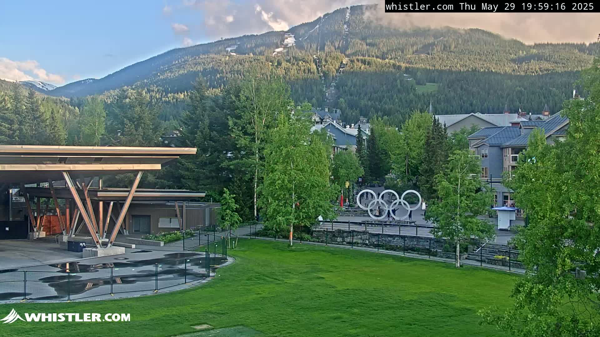 A partly cloudy afternoon scene shows a grassy area with a modern wooden structure and Olympic rings in the foreground, nestled in a village at the base of a mountain with a ski lift visible.