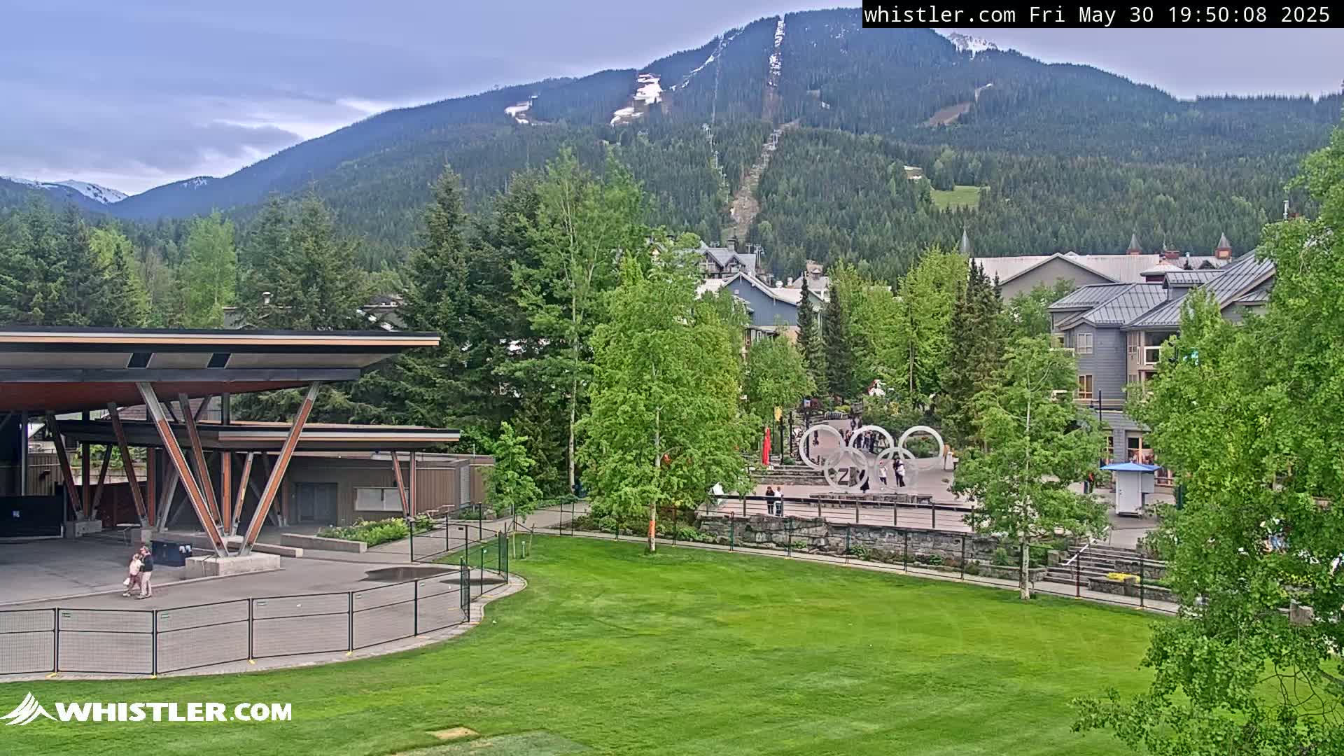 A partly cloudy day reveals a mountain village nestled in a valley, with a large grassy area in the foreground, a covered stage to the left, and the Olympic rings visible in the midground.