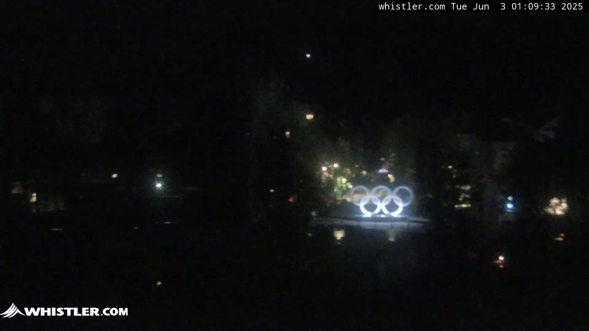 At night, the Olympic rings are illuminated near a dark body of water, with sparsely scattered lights visible in the surrounding area.