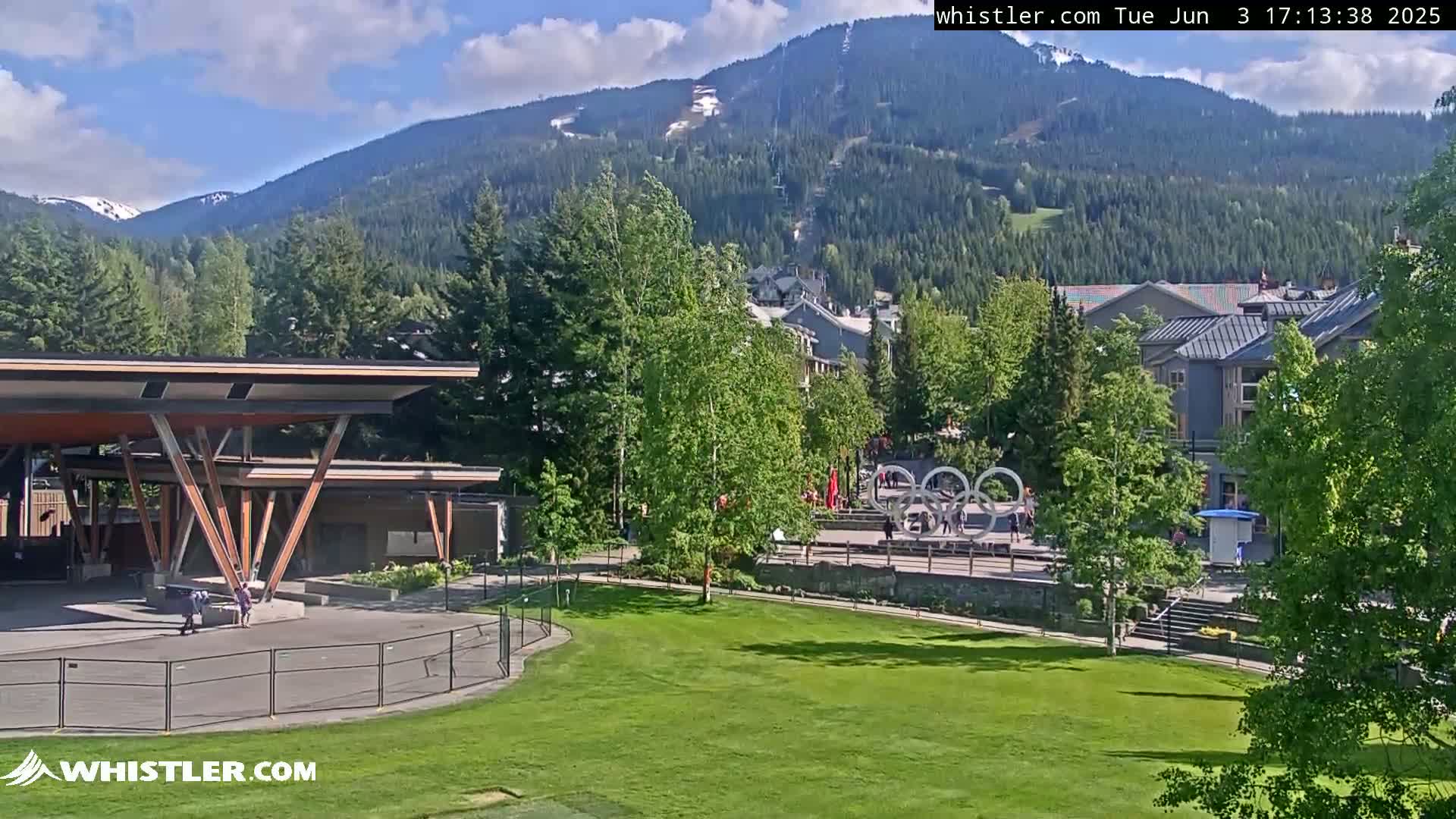 A grassy park area with a modern pavilion is situated below a village nestled in a valley, with mountains in the background under partly sunny skies.