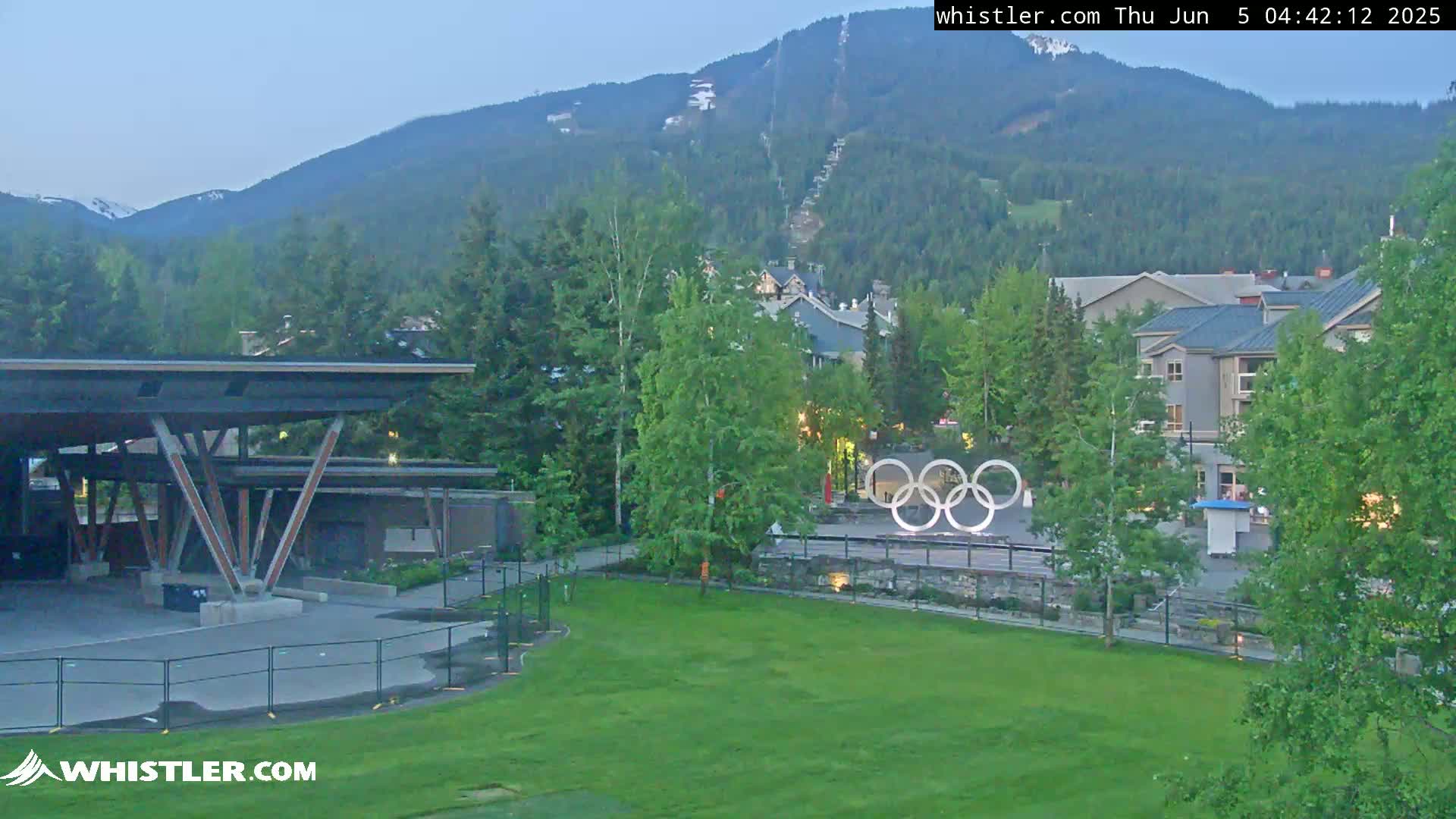 A mountain village scene at twilight shows a grassy area with an Olympic rings sculpture, buildings, trees, and a mountain with a ski lift in the background.