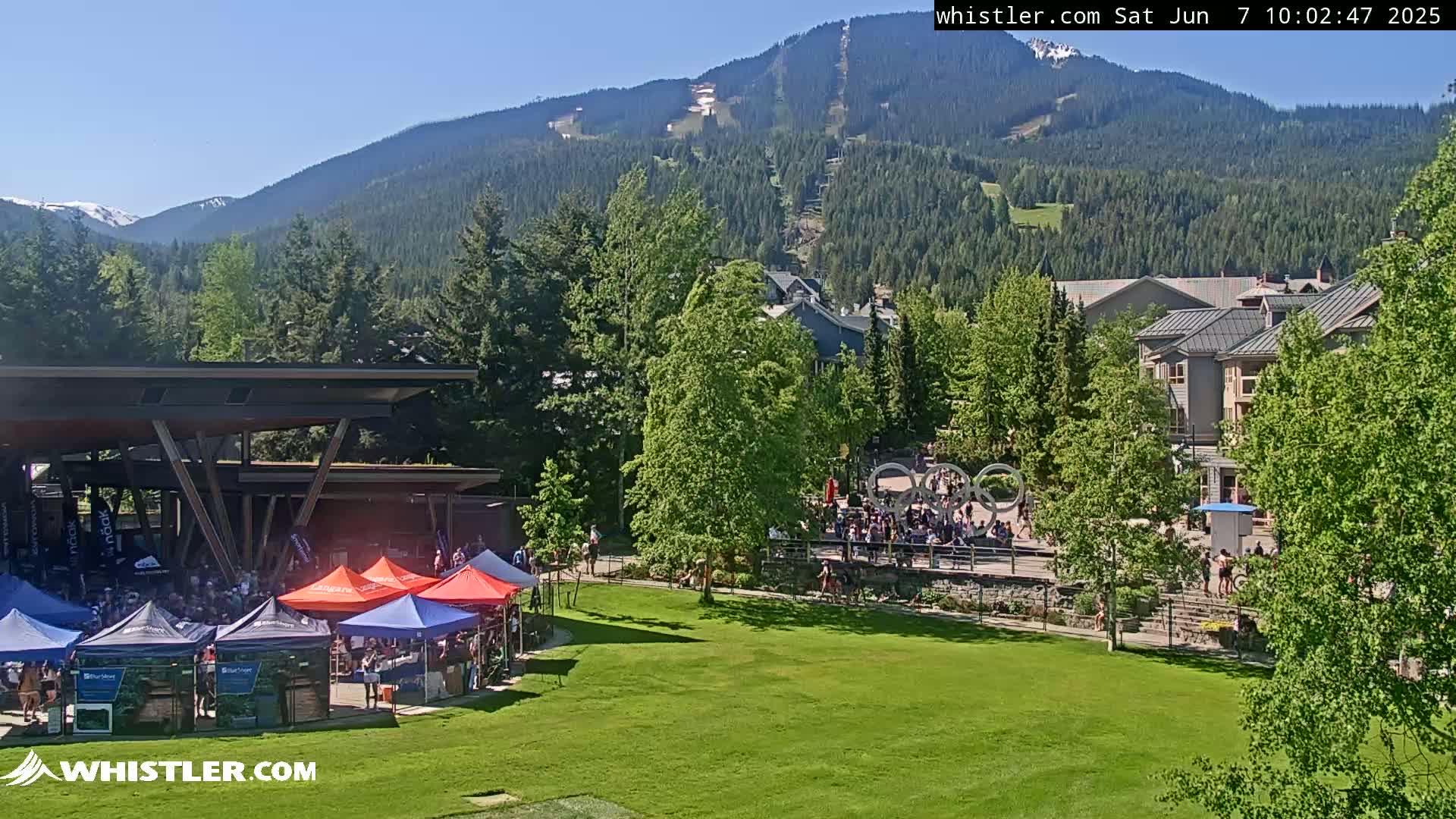 A sunny day reveals a mountainside ski resort with a village below, featuring a grassy area with several tents and people gathered near a covered structure.