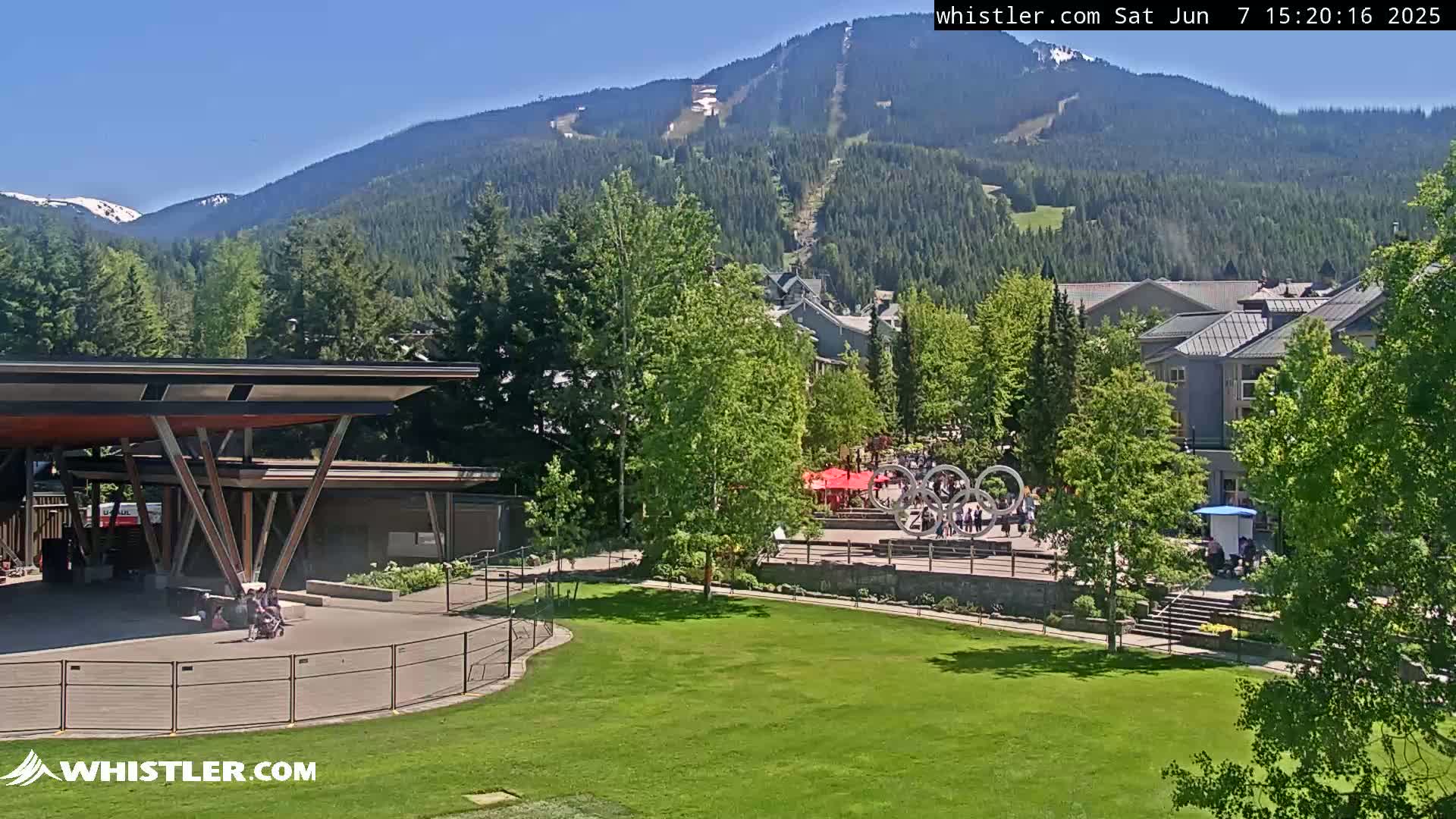 A sunny view of a grassy area, a modern covered structure, and buildings nestled in a green valley with mountains in the background, one of which shows a ski run.