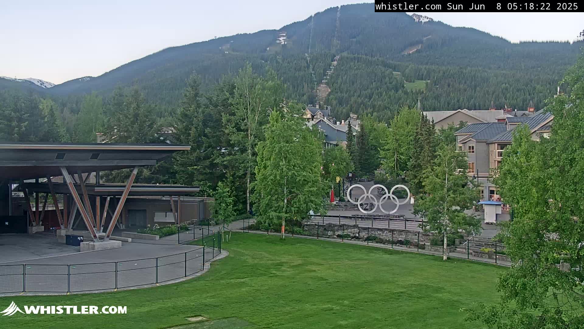 A grassy area with a modern pavilion and Olympic rings sits in front of a mountain village nestled against a mountainside under a clear sky.