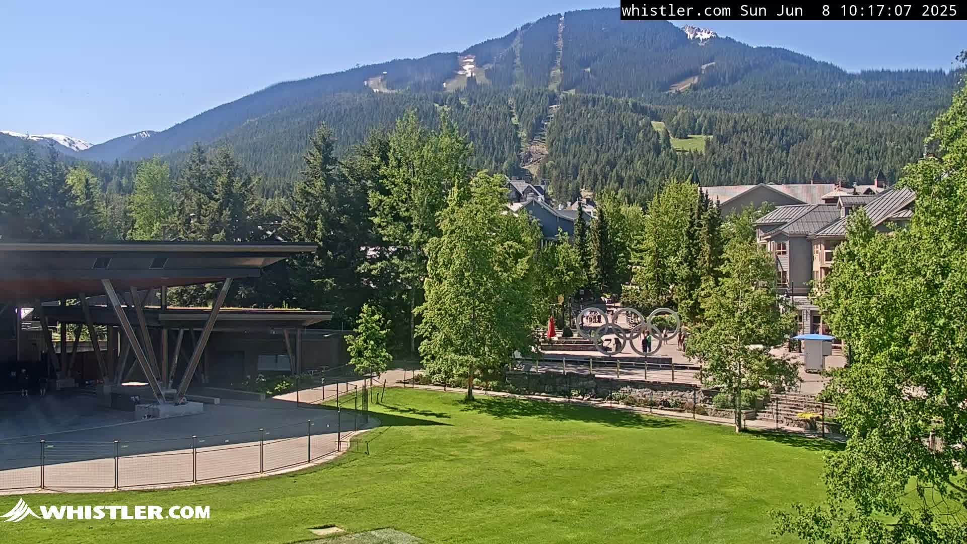 A sunny day reveals a mountainside ski resort with buildings, green lawns, and an outdoor pavilion nestled among lush trees.