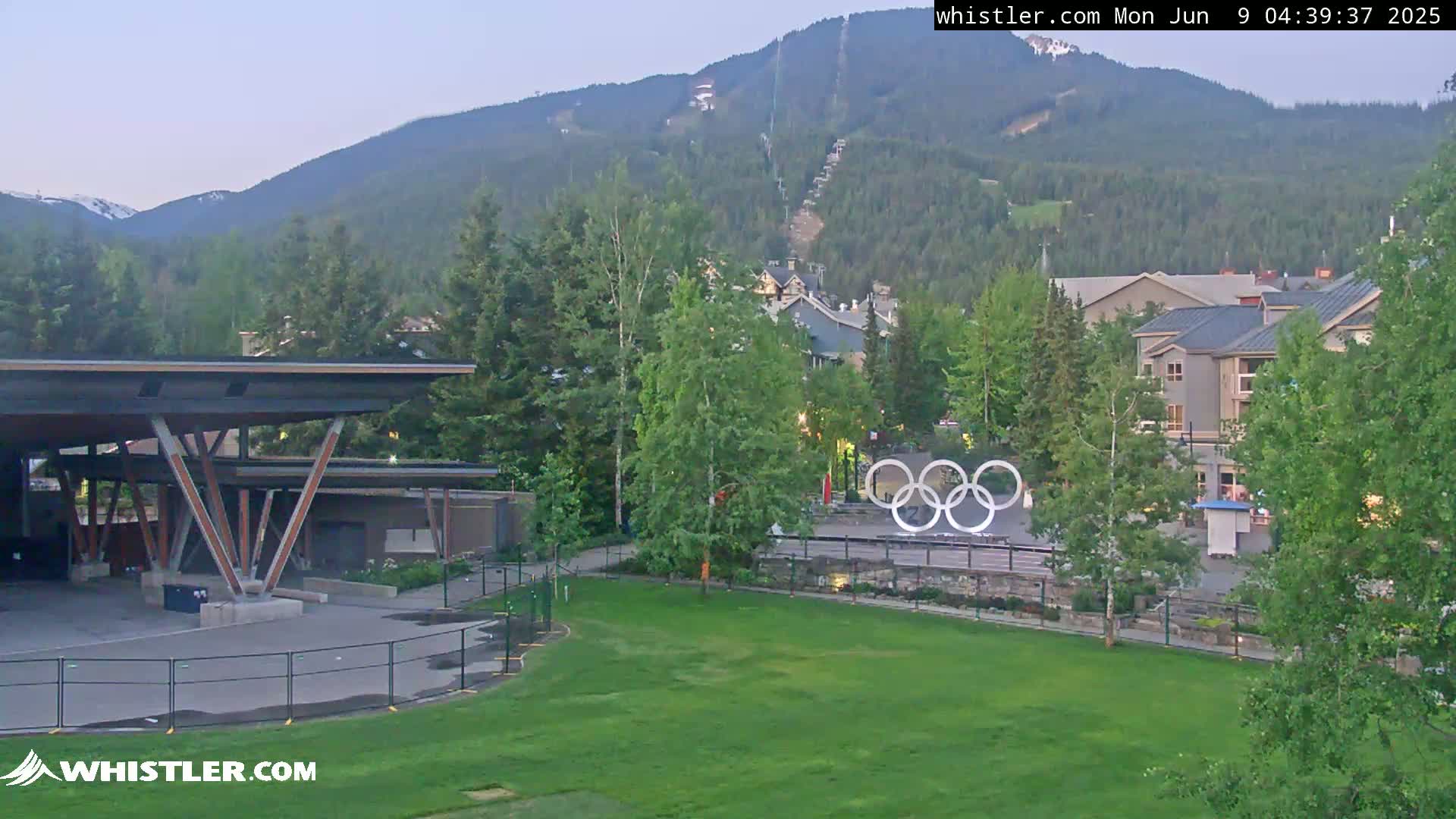 An outdoor view of a grassy area with an open-air pavilion, Olympic rings, buildings, and trees, all set against a backdrop of mountains under a clear, twilight sky.
