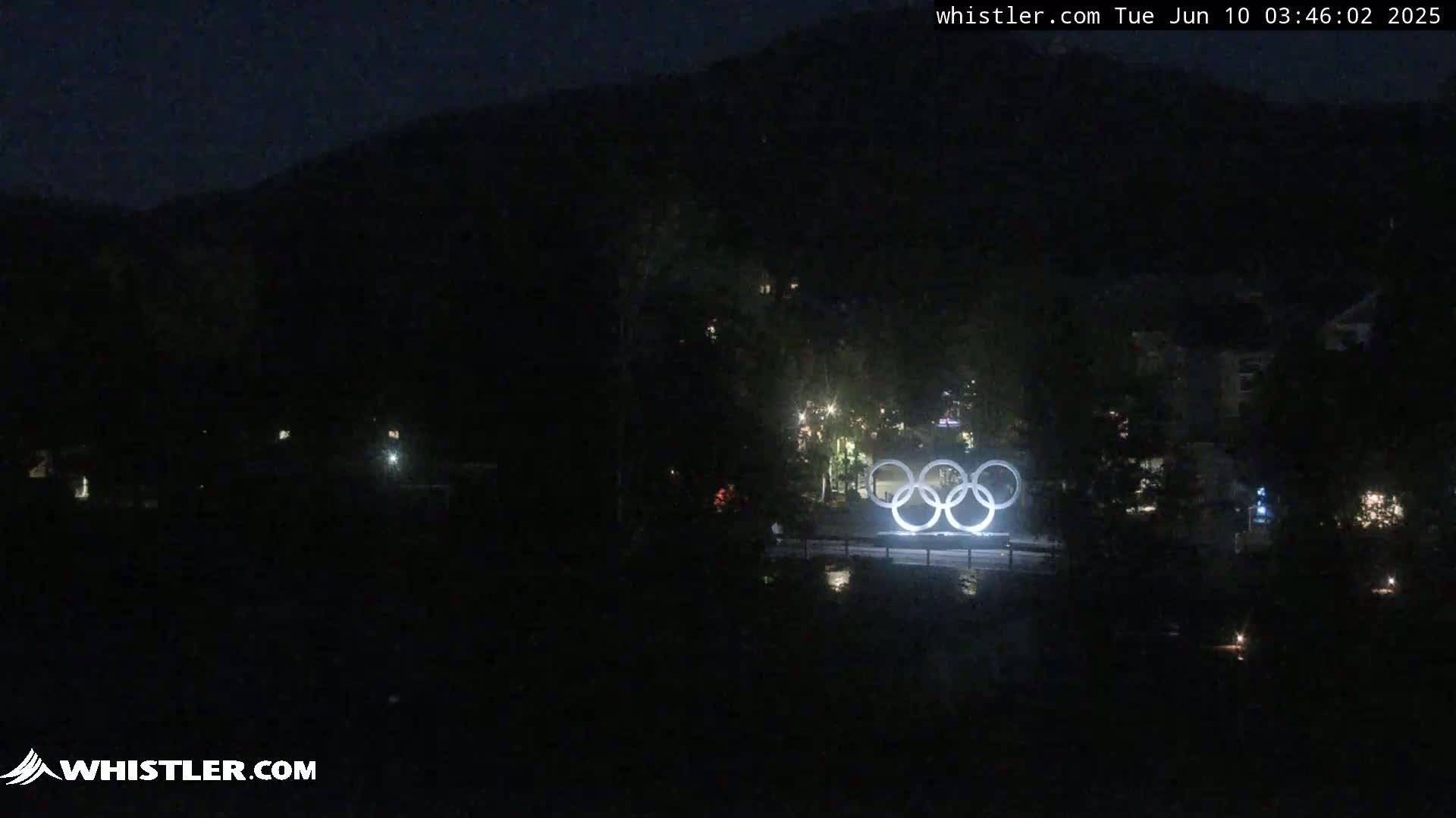 At night, the Olympic rings are illuminated in front of a dark mountain range under a clear sky.