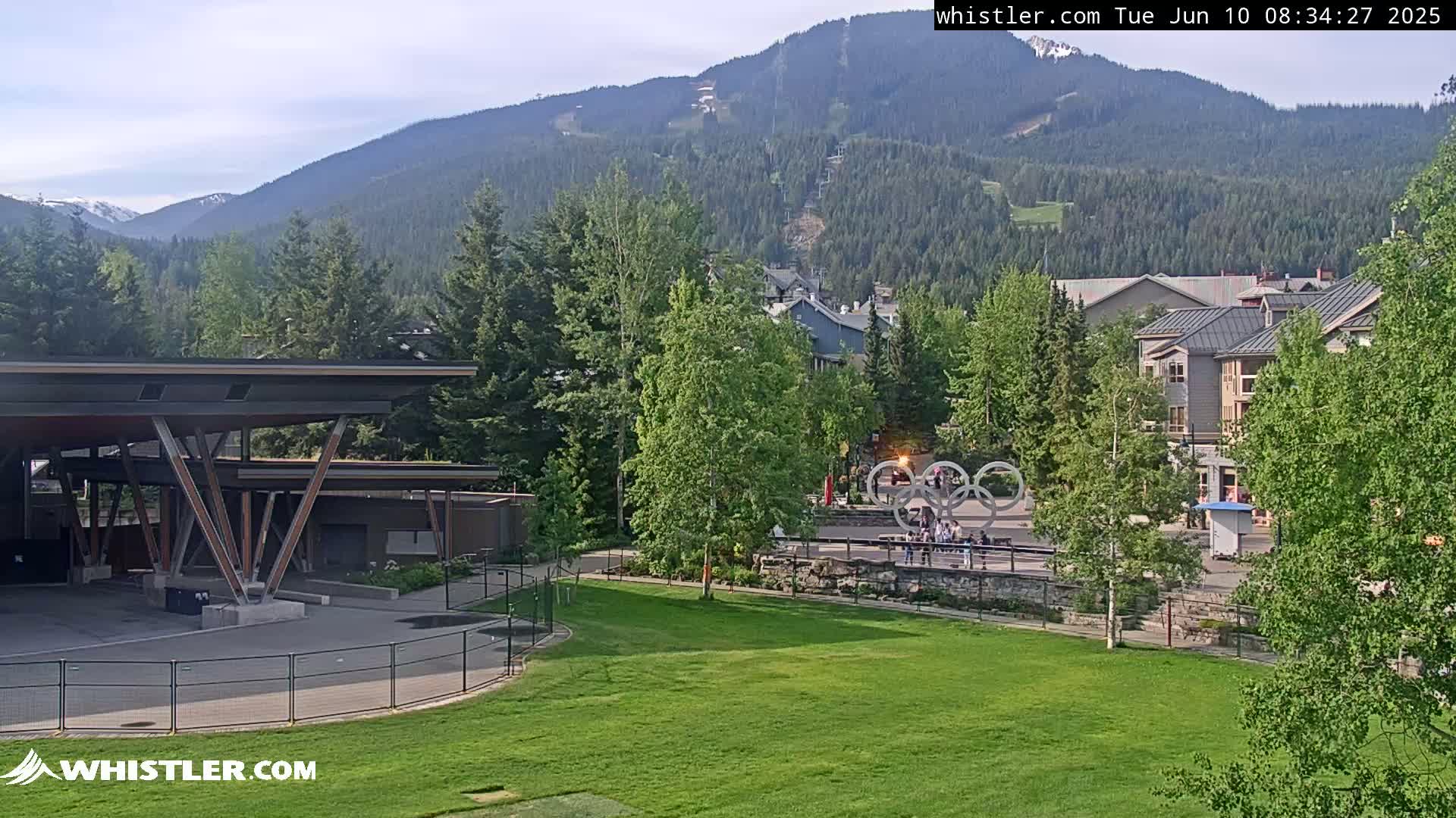 A grassy park with a modern building and Olympic rings sculpture is nestled amongst trees, with mountains in the background under a clear sky.