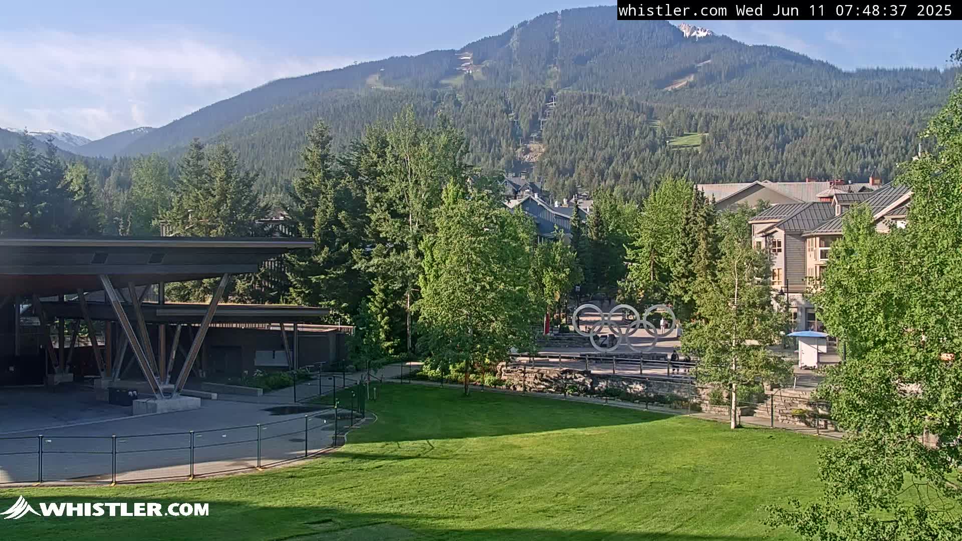 A grassy area with a modern pavilion and the Olympic rings is nestled in a village at the base of a mountain range under a mostly clear sky.