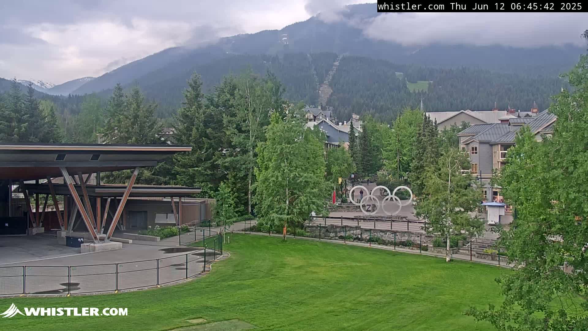 A mostly cloudy day reveals a mountainside ski lift, village buildings, and a grassy area with the Olympic rings, all viewed from an elevated vantage point near a covered pavilion.
