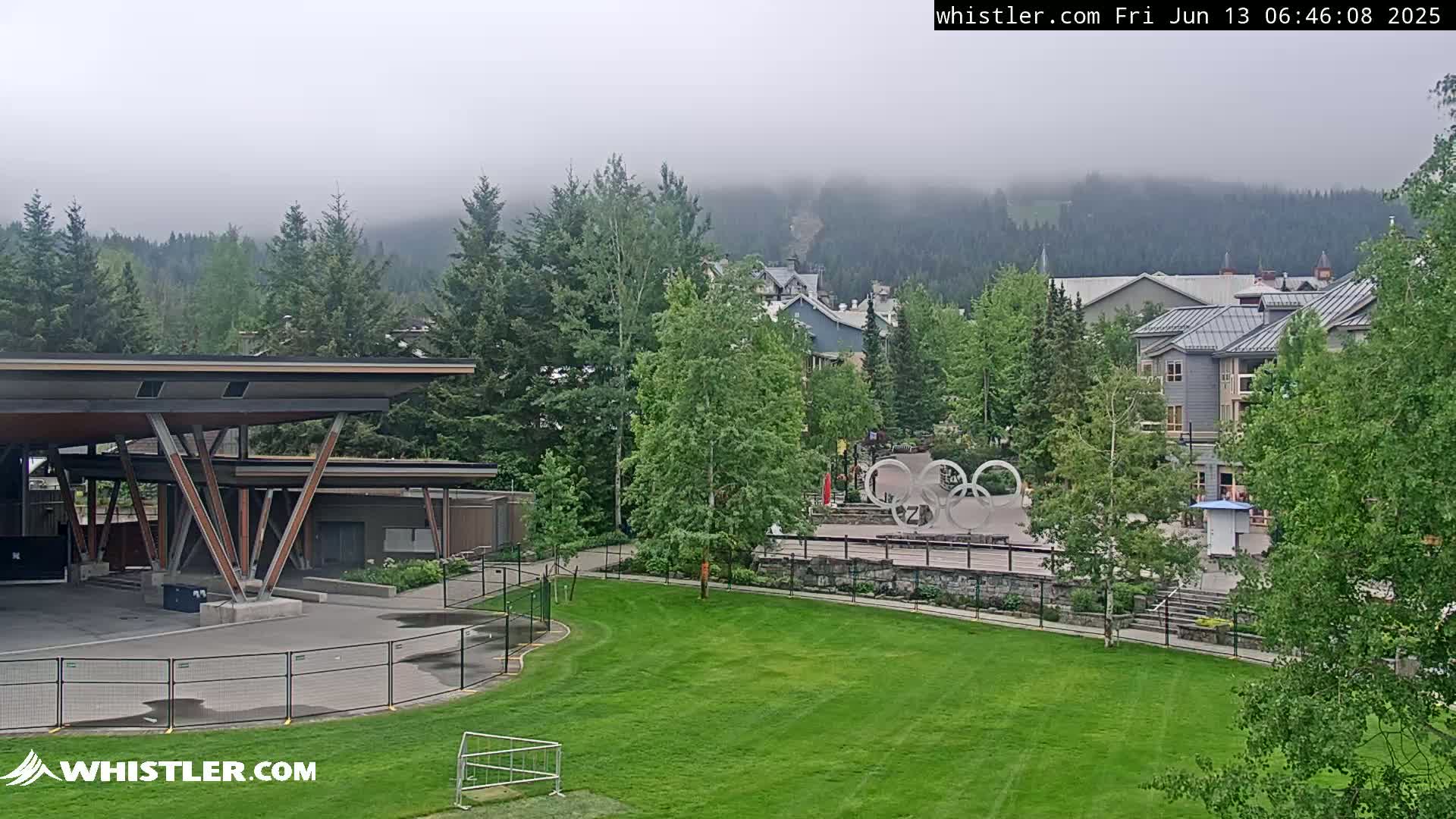 A grassy area with a covered structure and Olympic rings sits in front of buildings and a fog-covered mountain range.