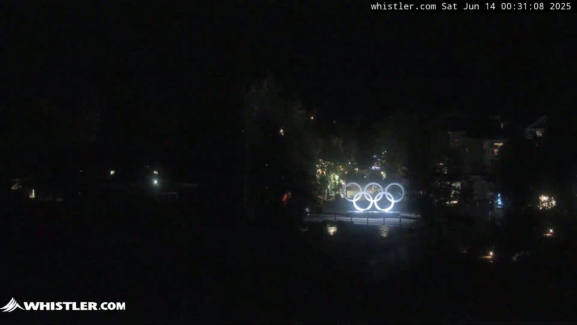 At night, illuminated Olympic rings are visible in the distance, surrounded by dark trees and buildings.
