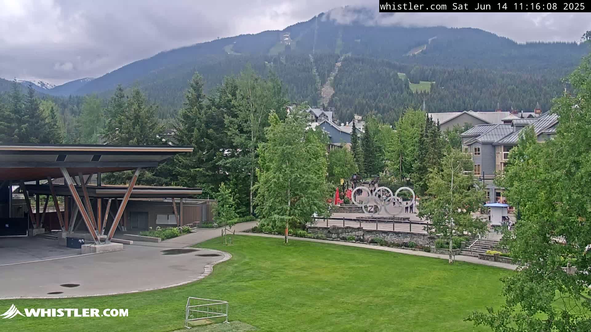 A grassy area with a modern covered structure is seen in the foreground, overlooking a village nestled amongst green trees and mountains under overcast skies.