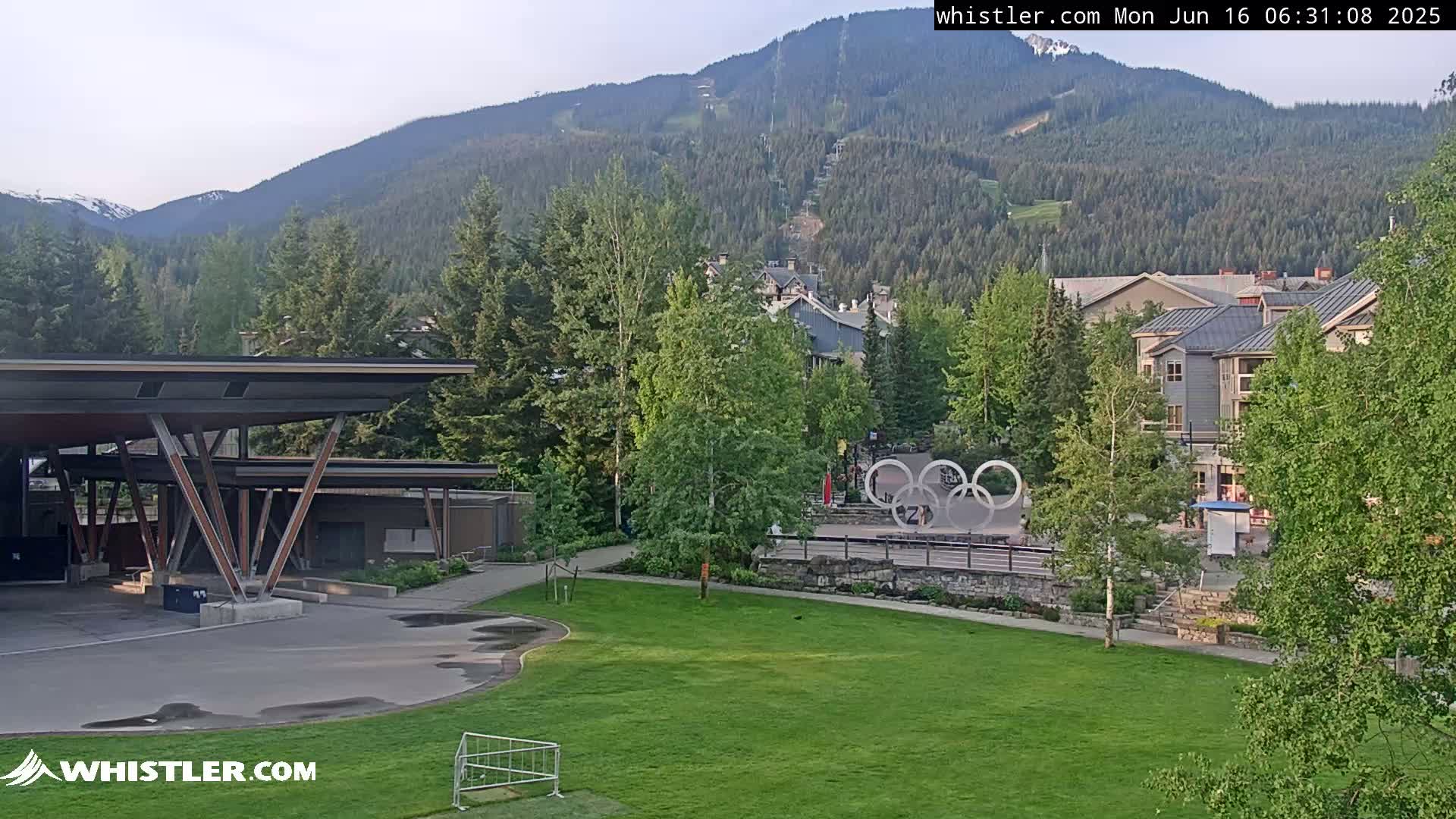A grassy area with a modern covered structure and Olympic rings sits in front of buildings and a mountain with a chairlift visible in the background under a mostly clear sky.