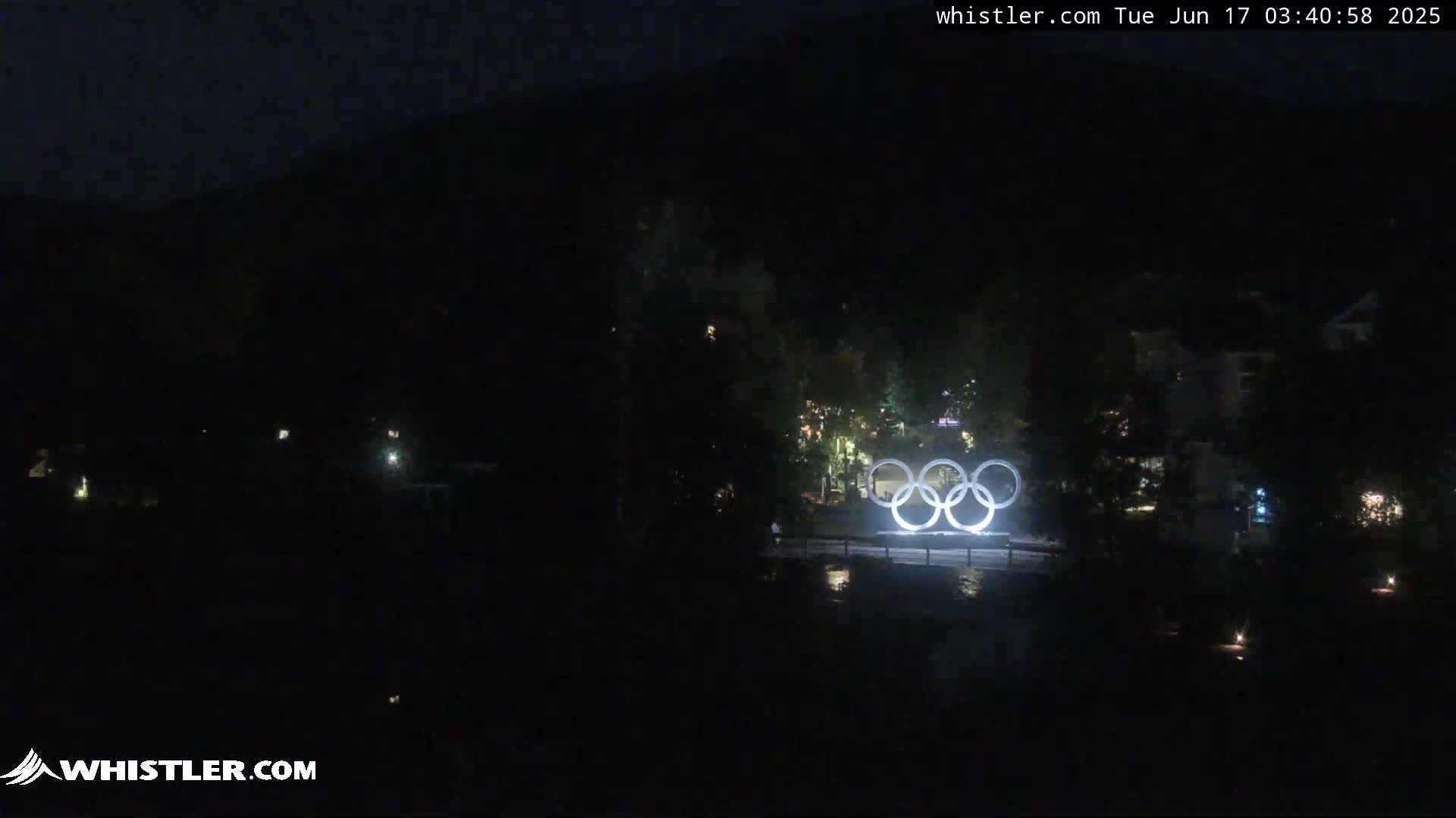 The Olympic rings are illuminated at night near a dark body of water, with a mountain in the background under a dark sky.