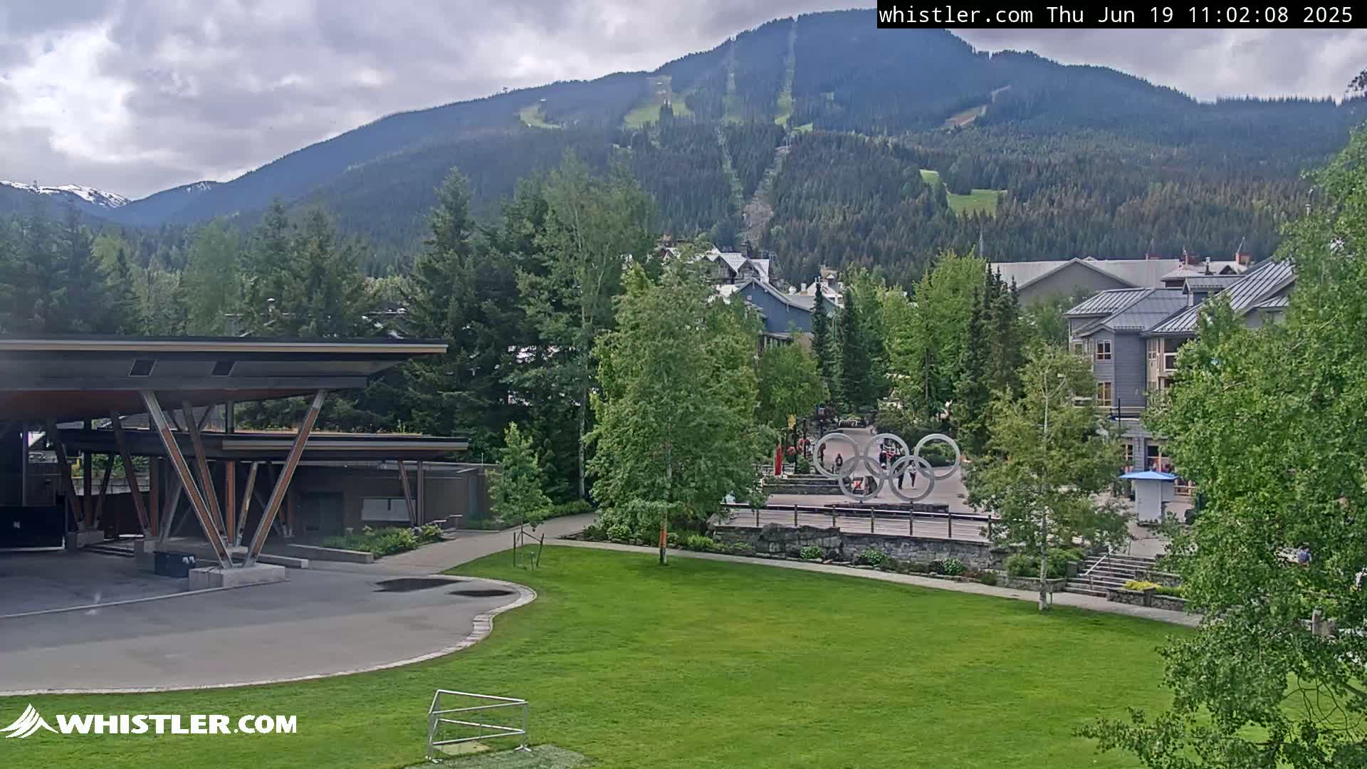 A view of a grassy park with a modern structure, Olympic rings, and buildings nestled in a valley below a mountain range under partly cloudy skies.