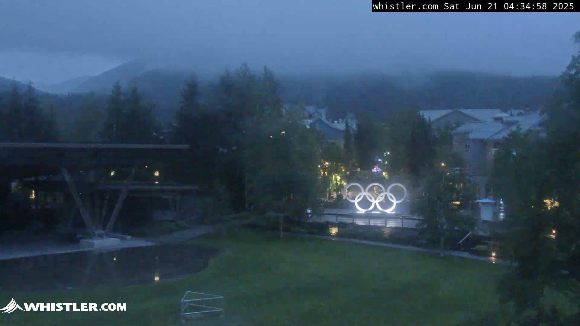 A dark, overcast scene shows the Olympic rings illuminated in a grassy area, with buildings and trees in the background under a gloomy sky.