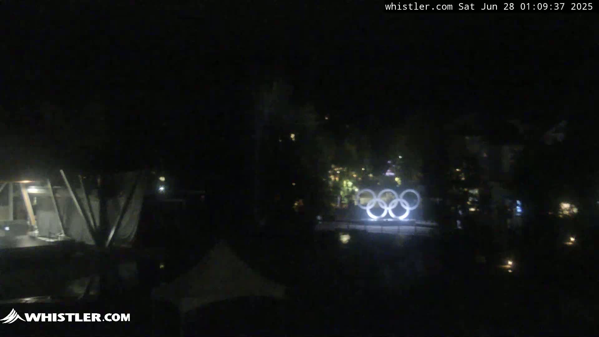 A nighttime, low-light view shows the Olympic rings illuminated in the distance, surrounded by dark trees and structures.