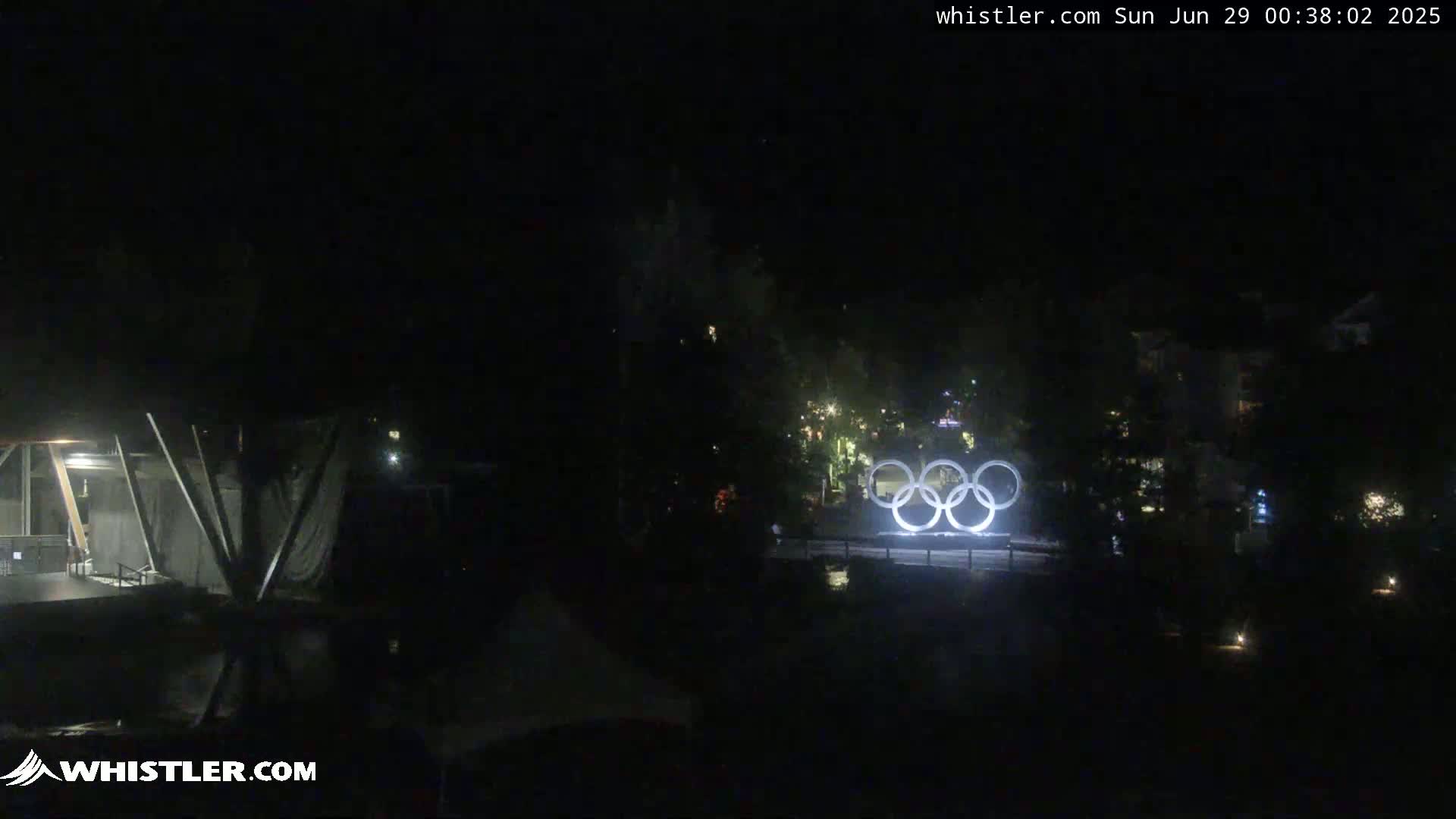 At night, illuminated Olympic rings are visible in the distance, with dark trees and buildings surrounding them.