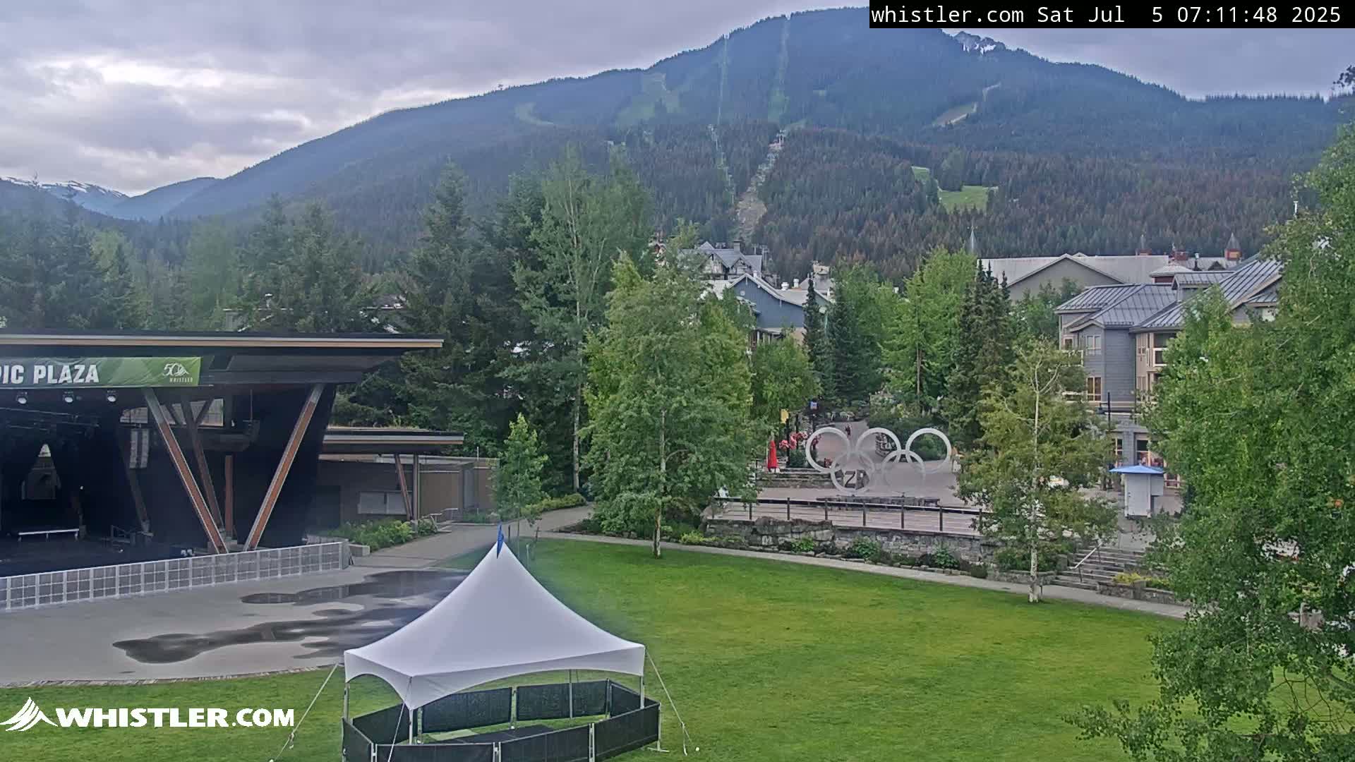 A grassy area with a white tent and an outdoor stage sits before a village nestled in a valley beneath a mountain range on an overcast day.