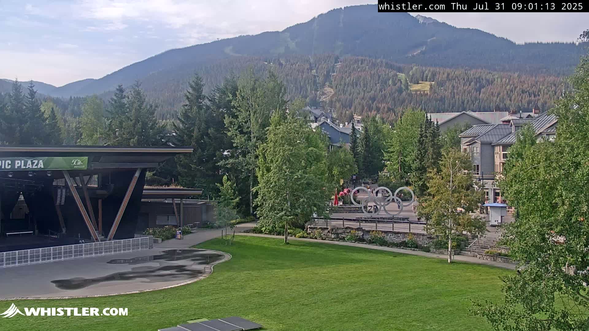 A mostly sunny day reveals a mountainside village with an outdoor stage, Olympic rings, and lush green lawns under partly cloudy skies.