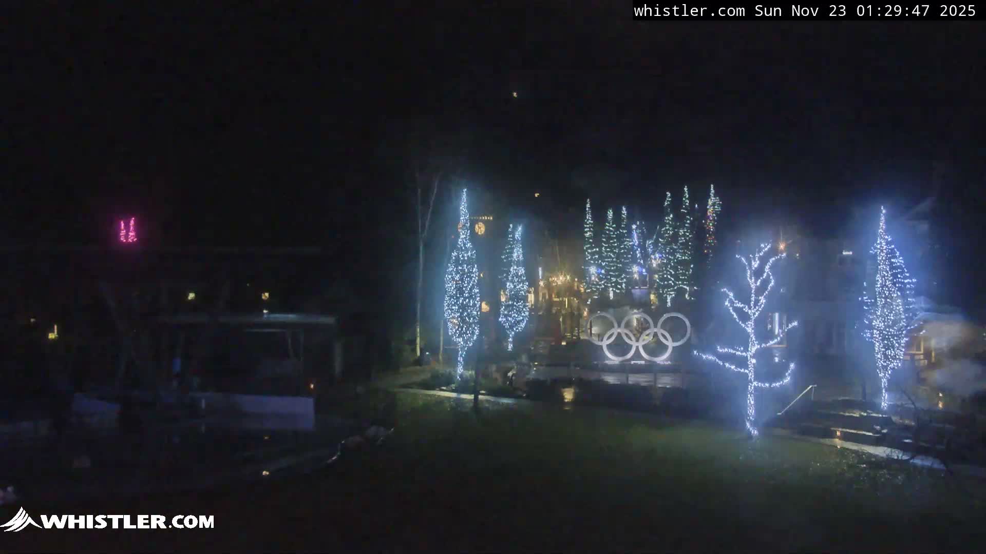 A festive outdoor plaza is illuminated at night with numerous trees covered in bright white string lights and a large, glowing Olympic Rings sculpture, all under clear and calm weather conditions.
