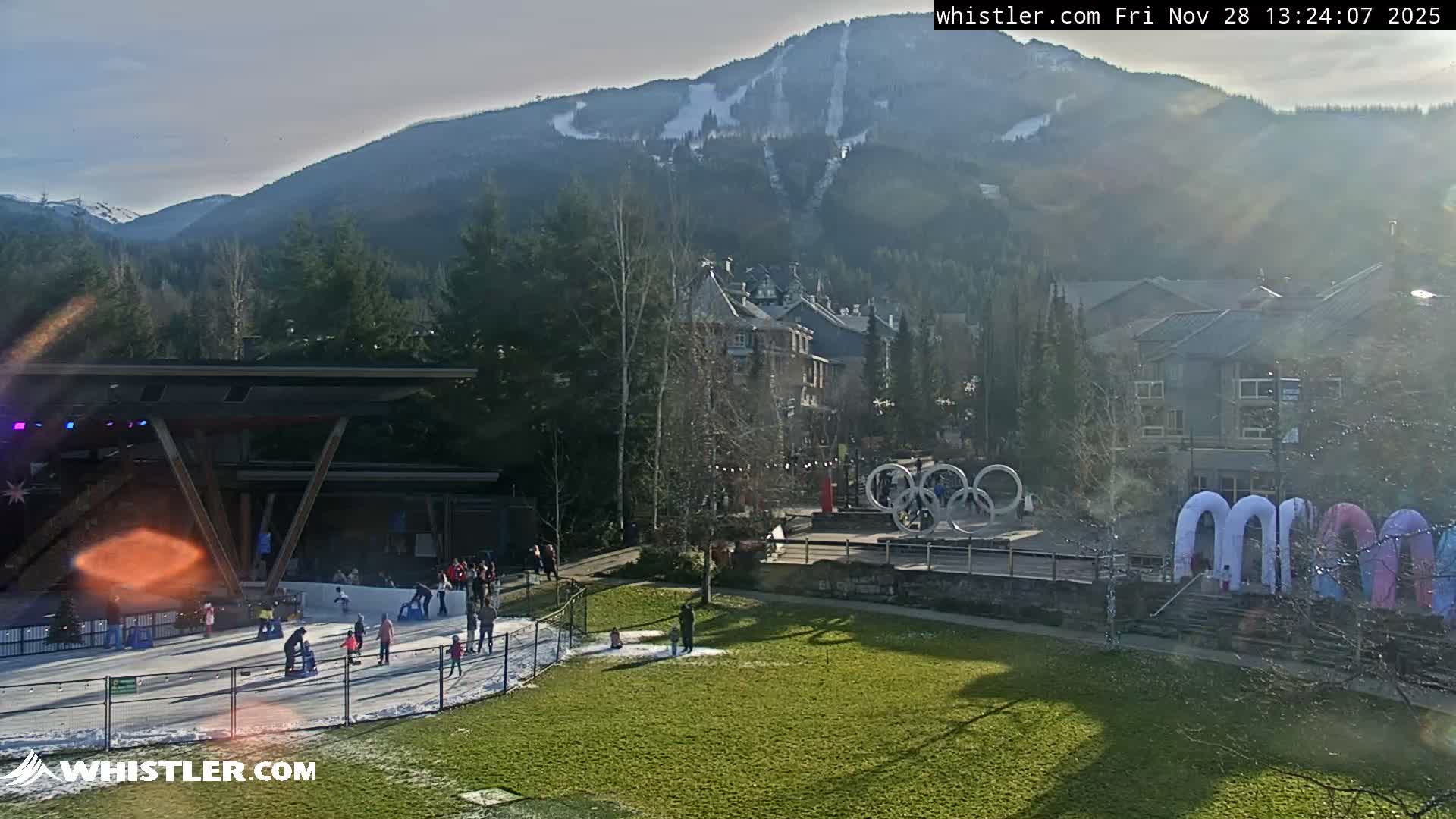 An outdoor scene depicts people ice skating on a rink beside a green grassy area with patches of snow, set against a backdrop of buildings, evergreen trees, and snow-dusted mountains under a partly cloudy and sunny sky.