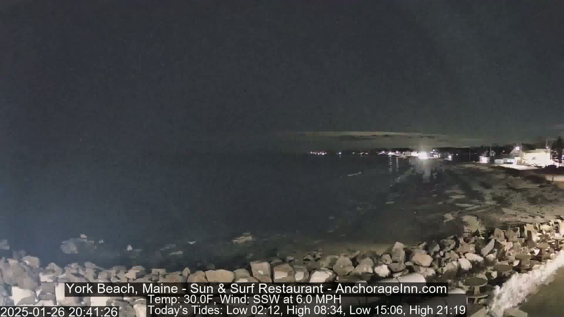 A nighttime, coastal scene shows a dark, rocky shoreline meeting a calm, dark body of water under a clear, dark sky.