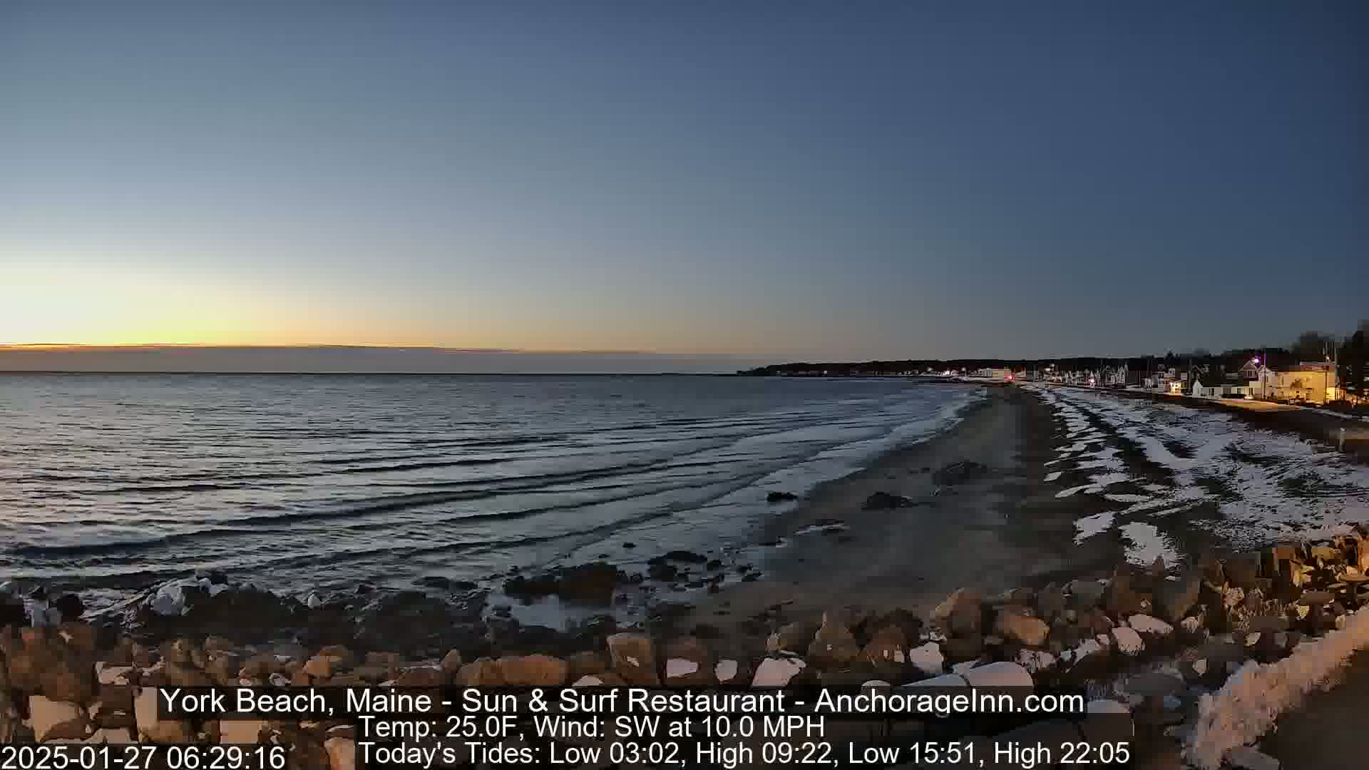 A twilight beach scene shows a calm ocean, a sandy shore partially covered in snow, and a town in the distance under a clear sky.