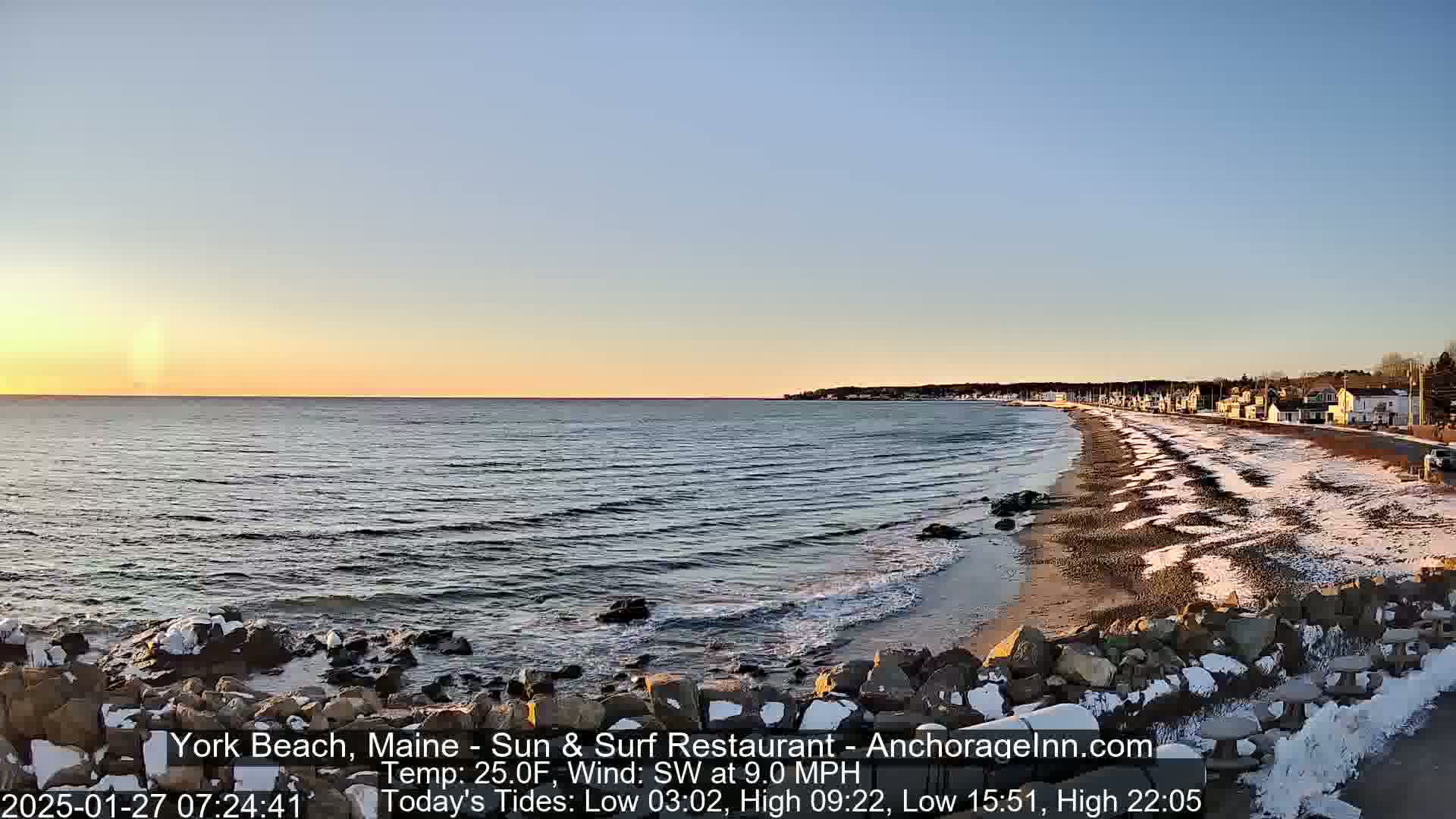 A calm ocean meets a snow-dusted beach at sunset, with a small town visible in the distance.