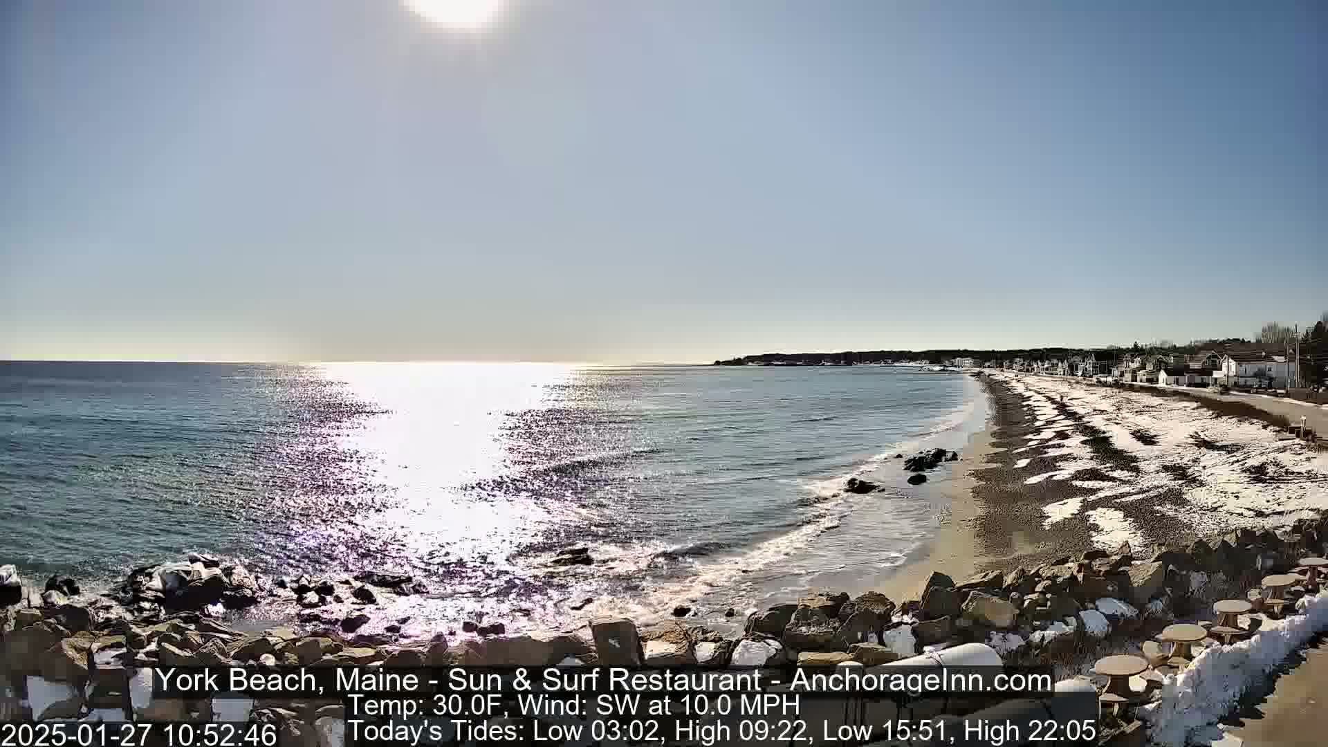A sunny, partly snowy beach with calm ocean waves and a shoreline lined with houses is visible from an elevated vantage point.